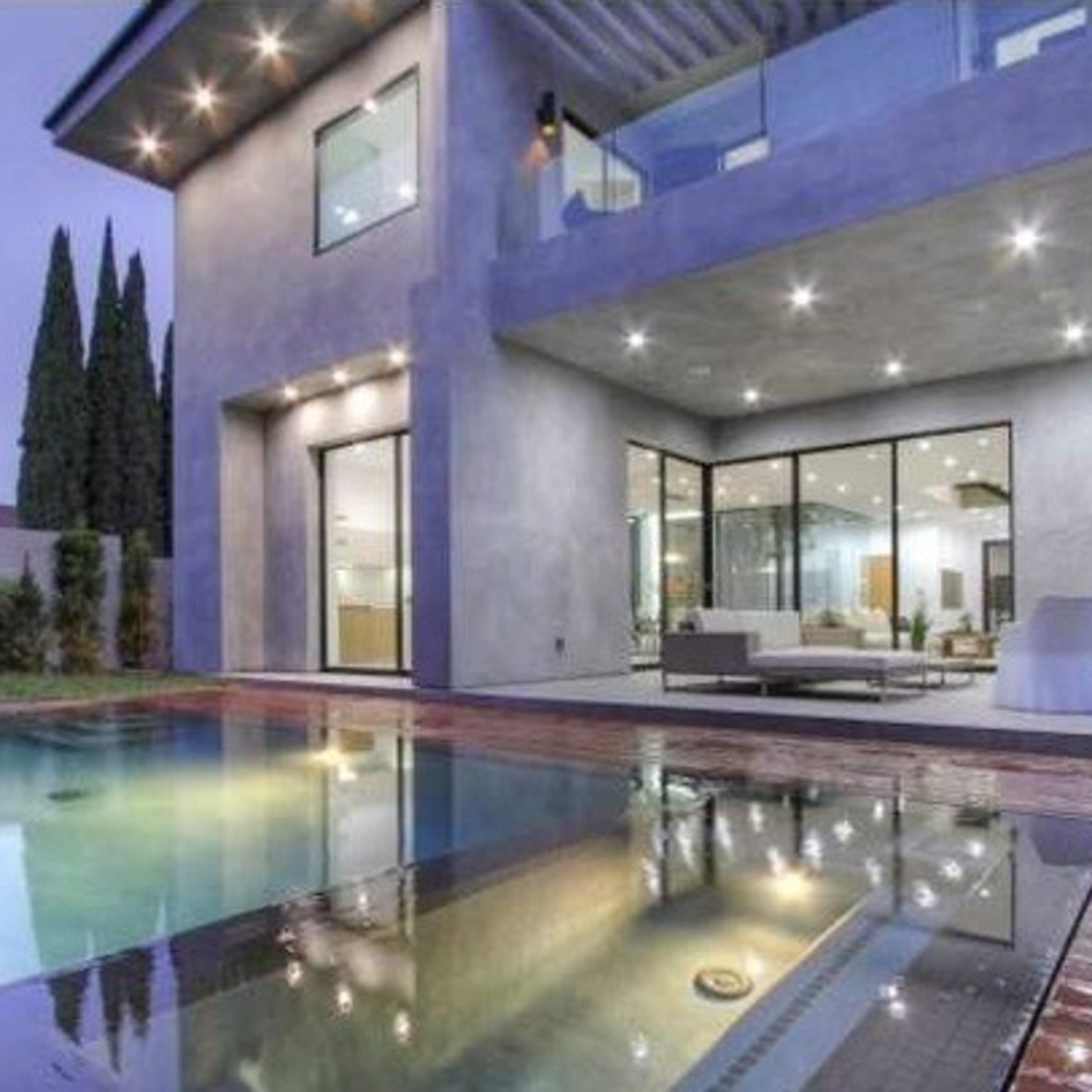 A modern house features a pool in the foreground and large glass windows showcasing a living area in the background.
