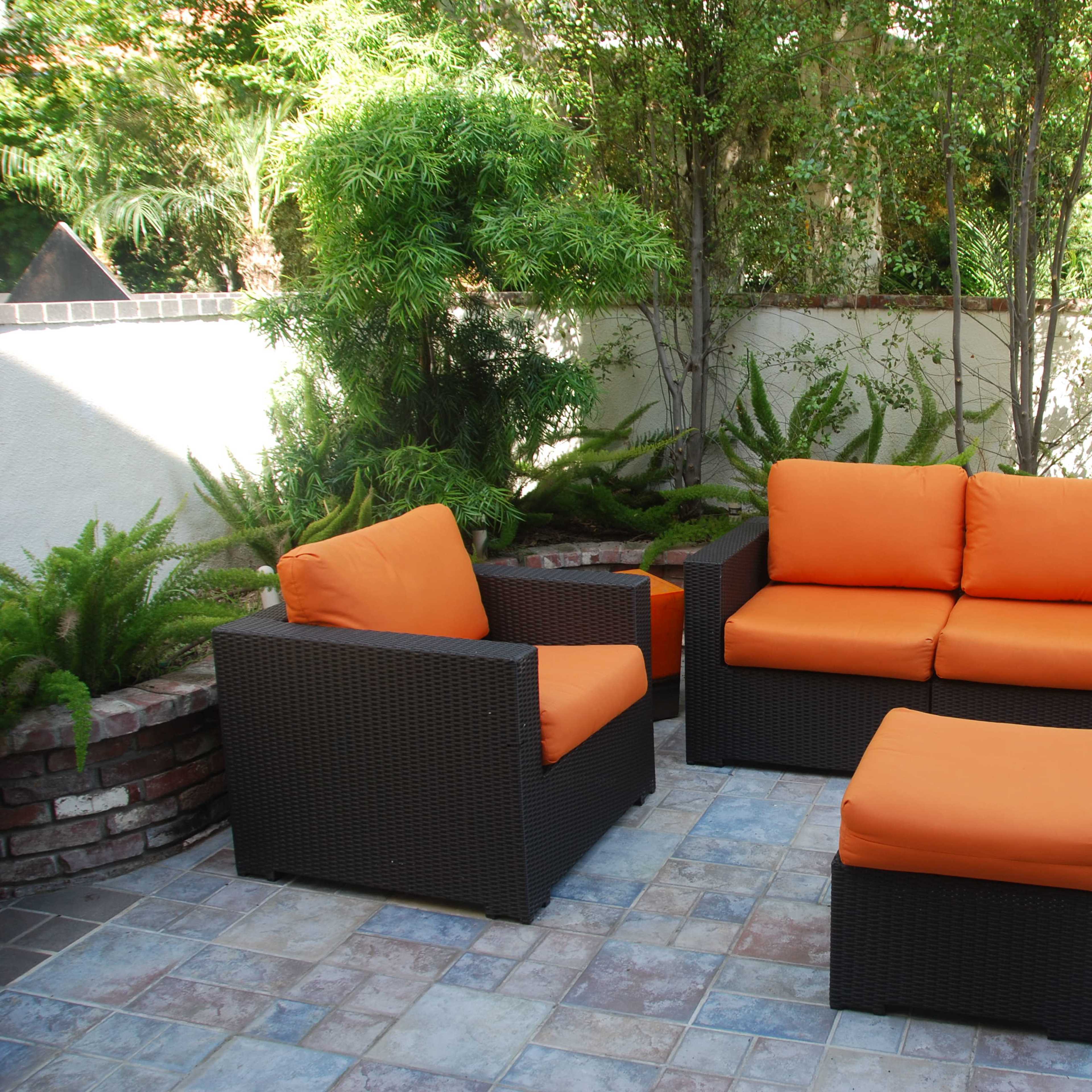 A patio area features a dark wicker seating set with orange cushions, accompanied by a matching ottoman, surrounded by greenery.