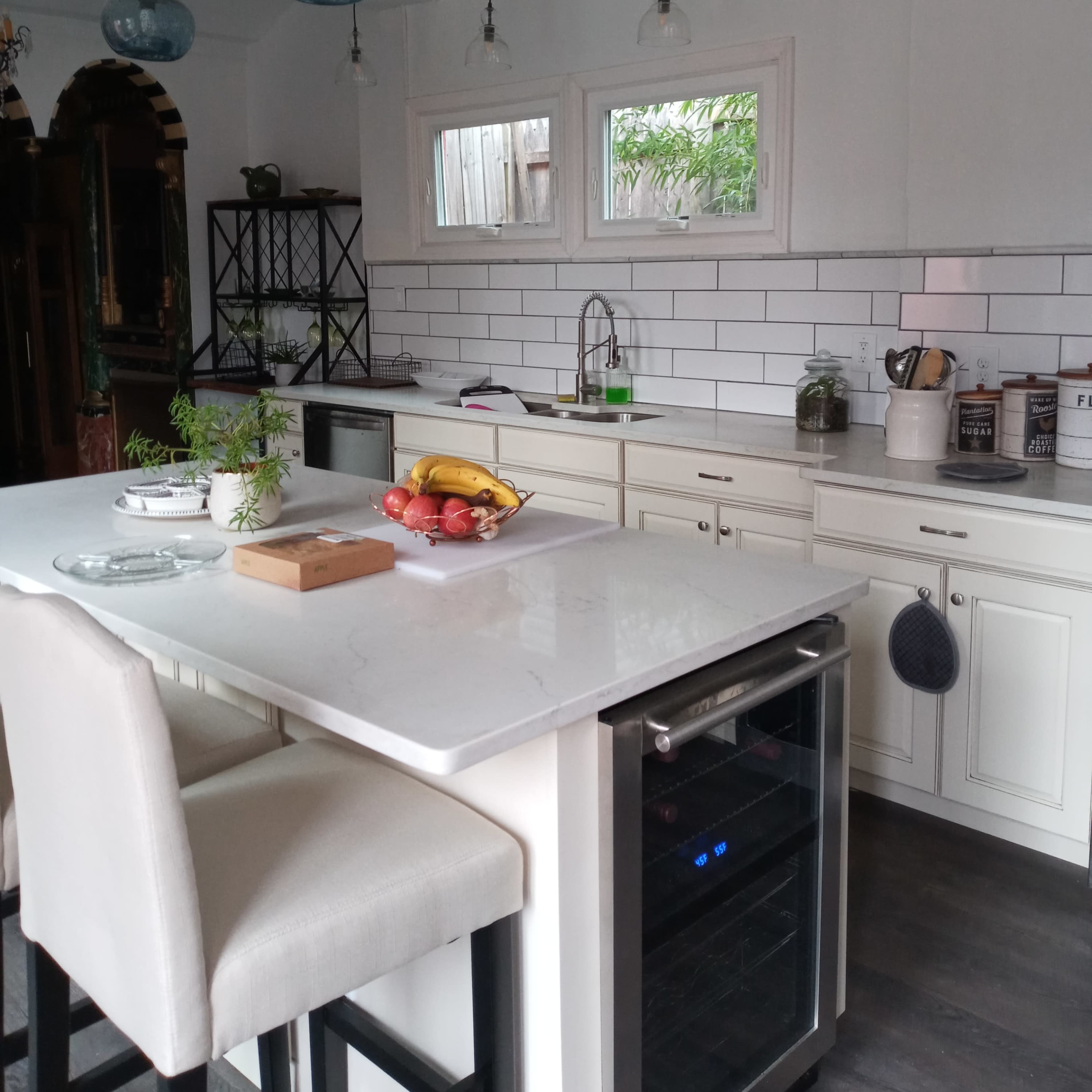 A modern kitchen features a white island with bar stools, pendant lights hanging from a vaulted ceiling, and various fruits arranged on the counter.