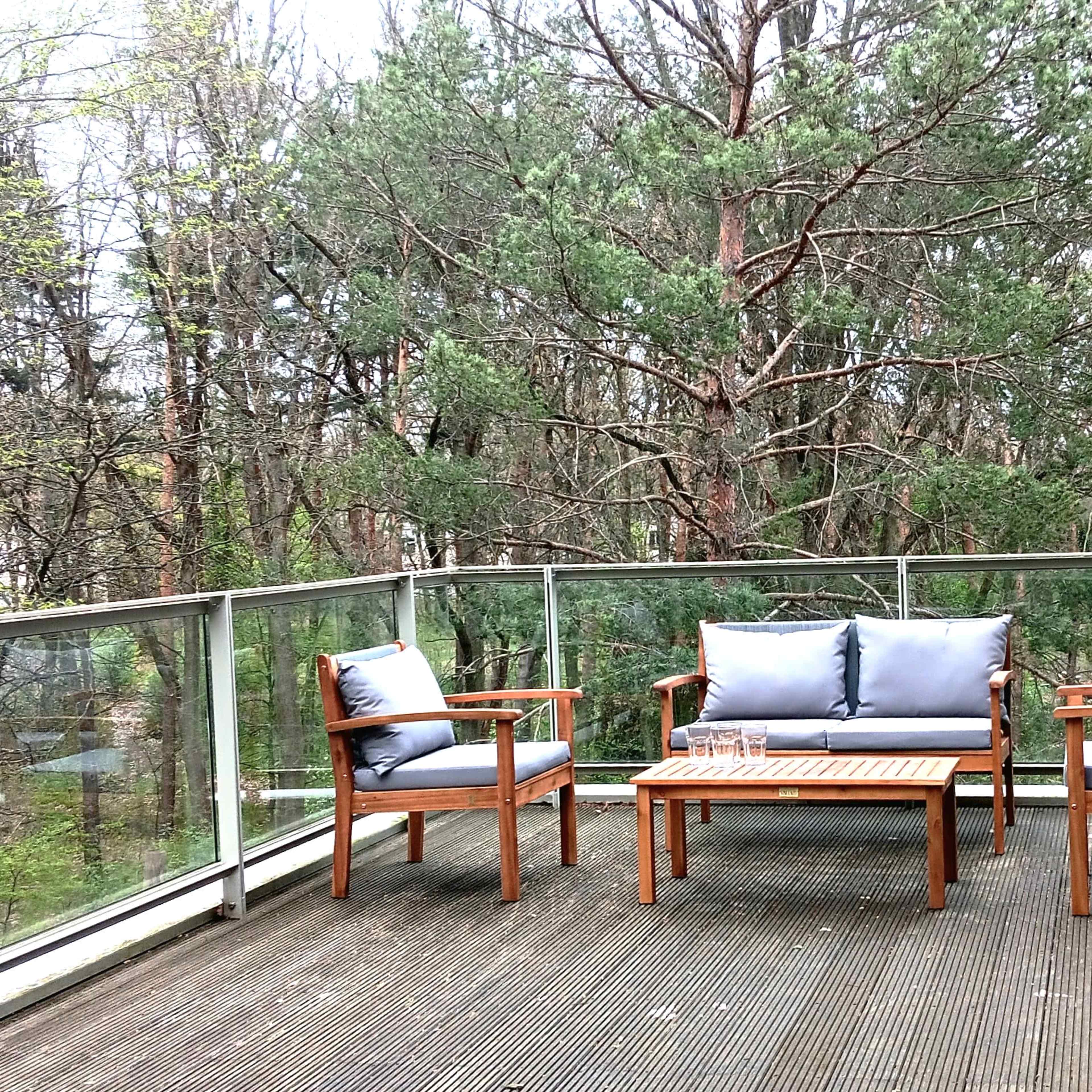 A balcony features wooden furniture with cushions, overlooking a forested area.
