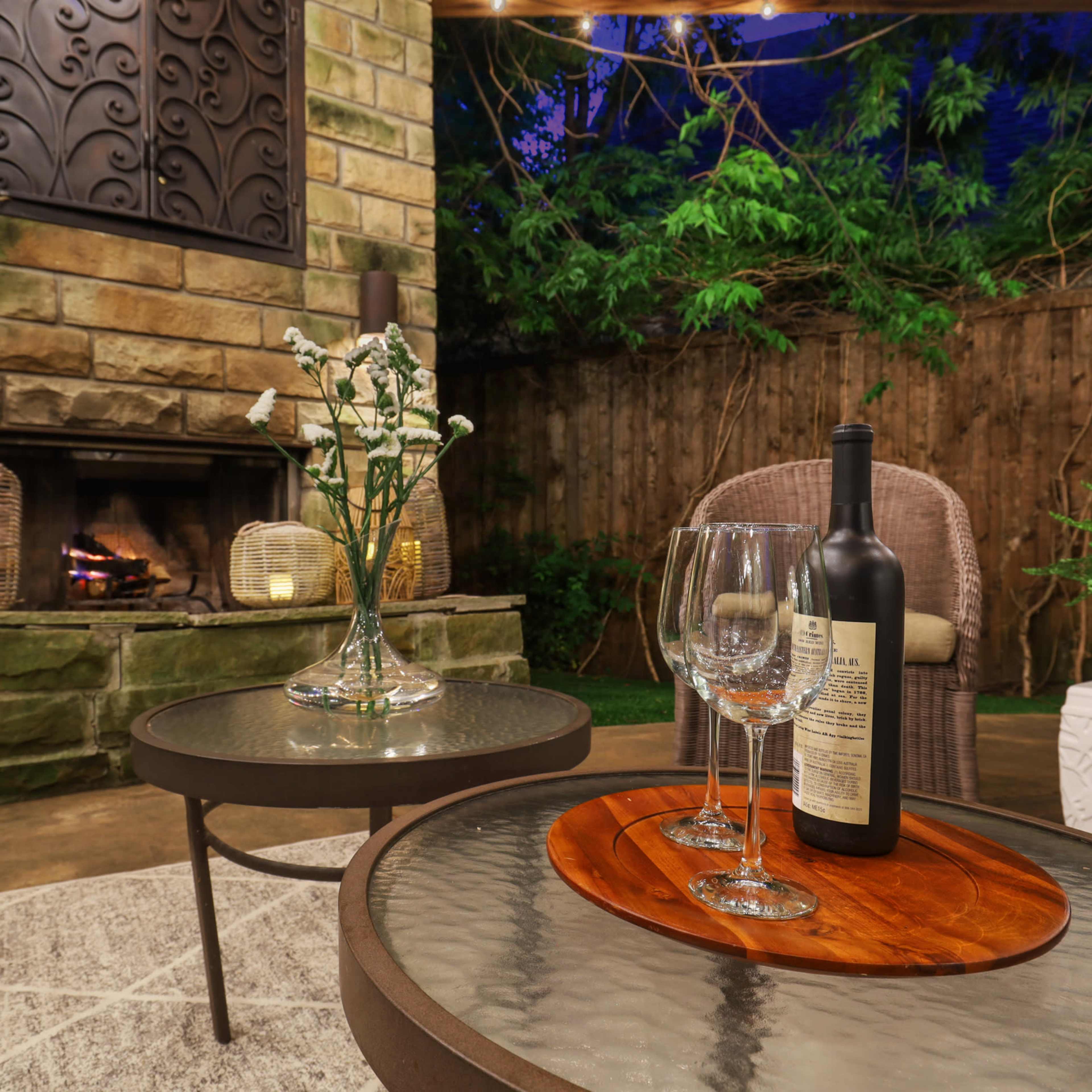 A cozy outdoor seating area features a stone fireplace, a glass coffee table with a wooden serving tray and wine bottle, and lush greenery in the background.