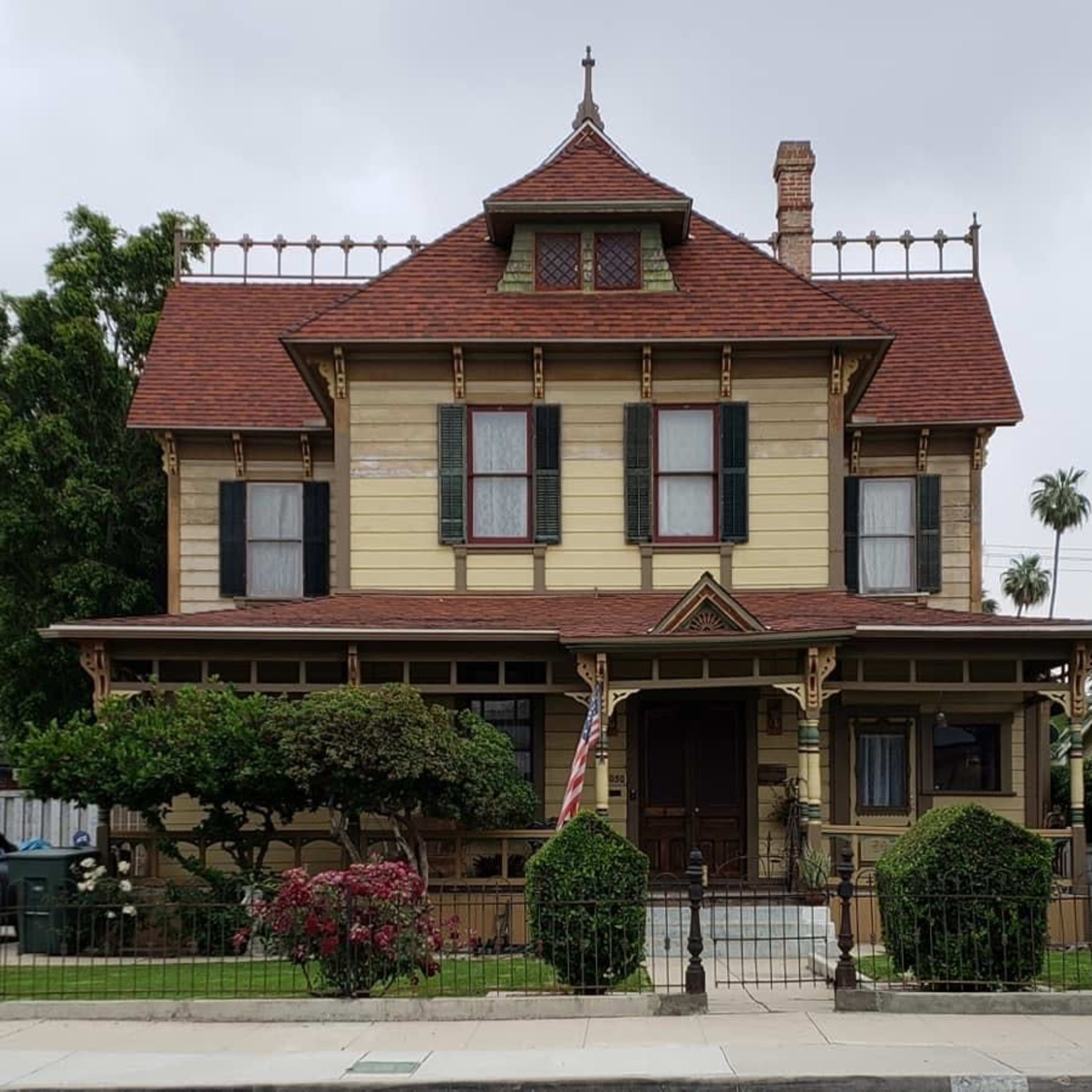 A Victorian-style house with a multi-gabled roof, ornate detailing, and a front porch surrounded by manicured bushes and a railing.