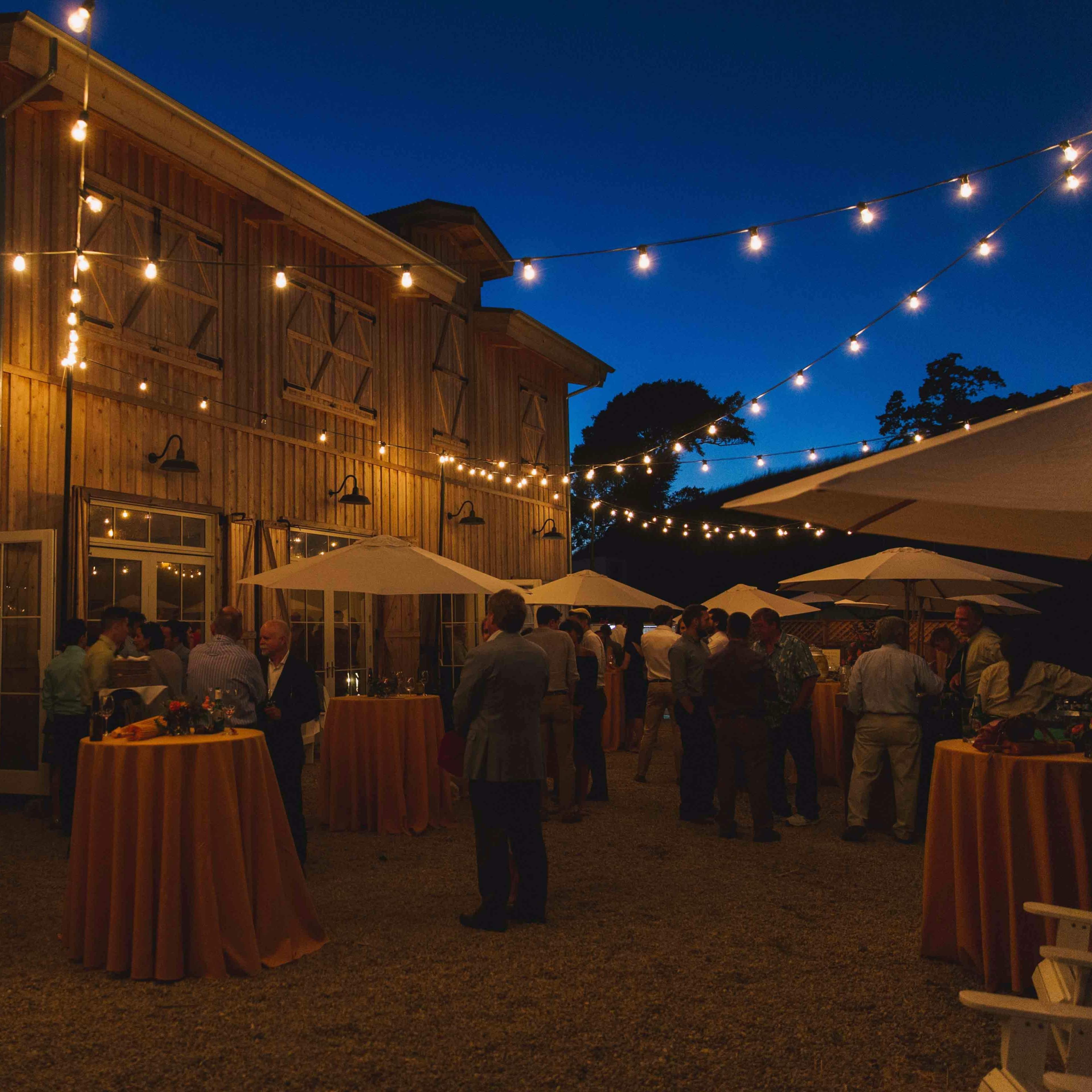 A gathering of guests is taking place outdoors near a barn, illuminated by string lights and accompanied by tables with umbrellas.