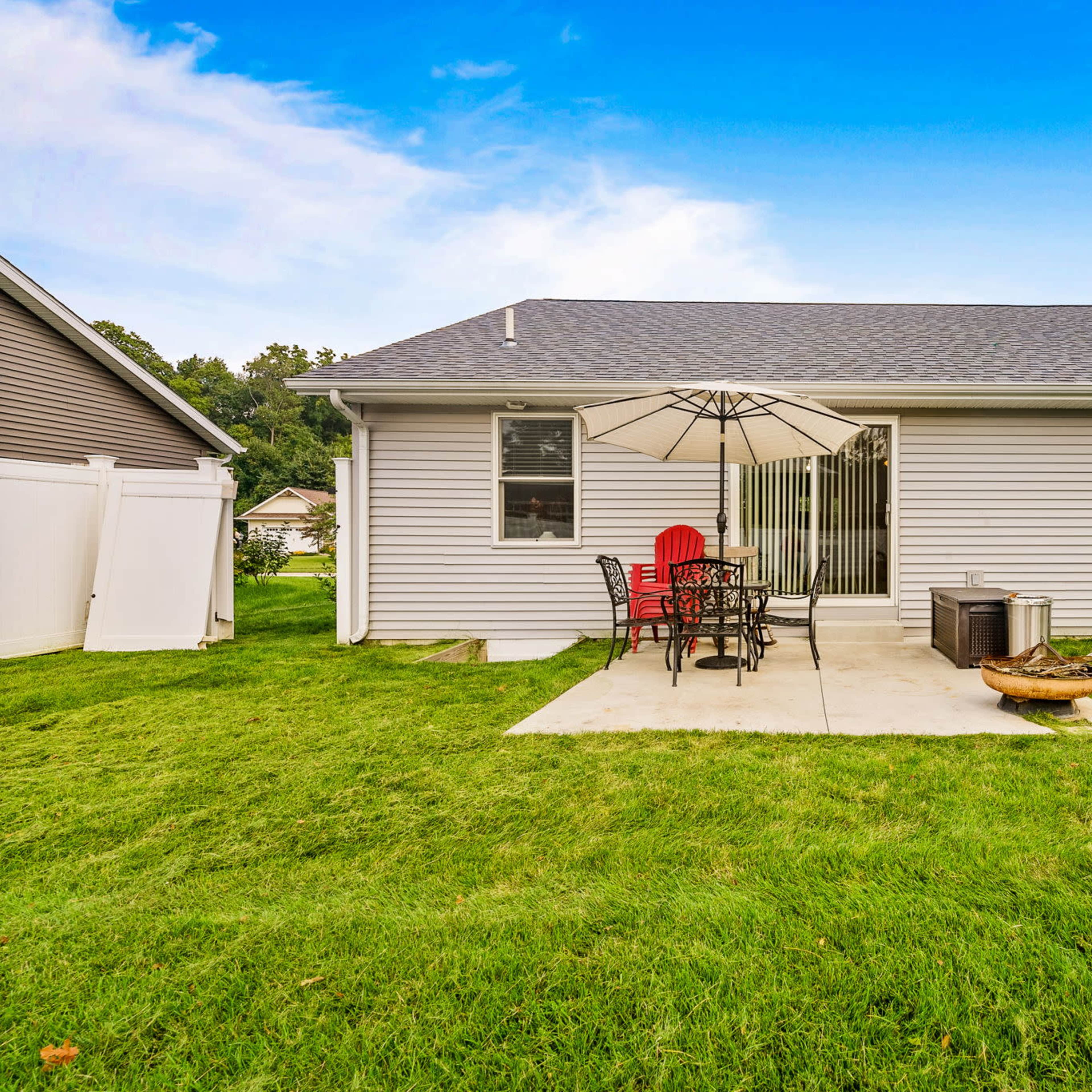 A backyard patio with a table and chairs under an umbrella, surrounded by a lawn and adjacent to a house.