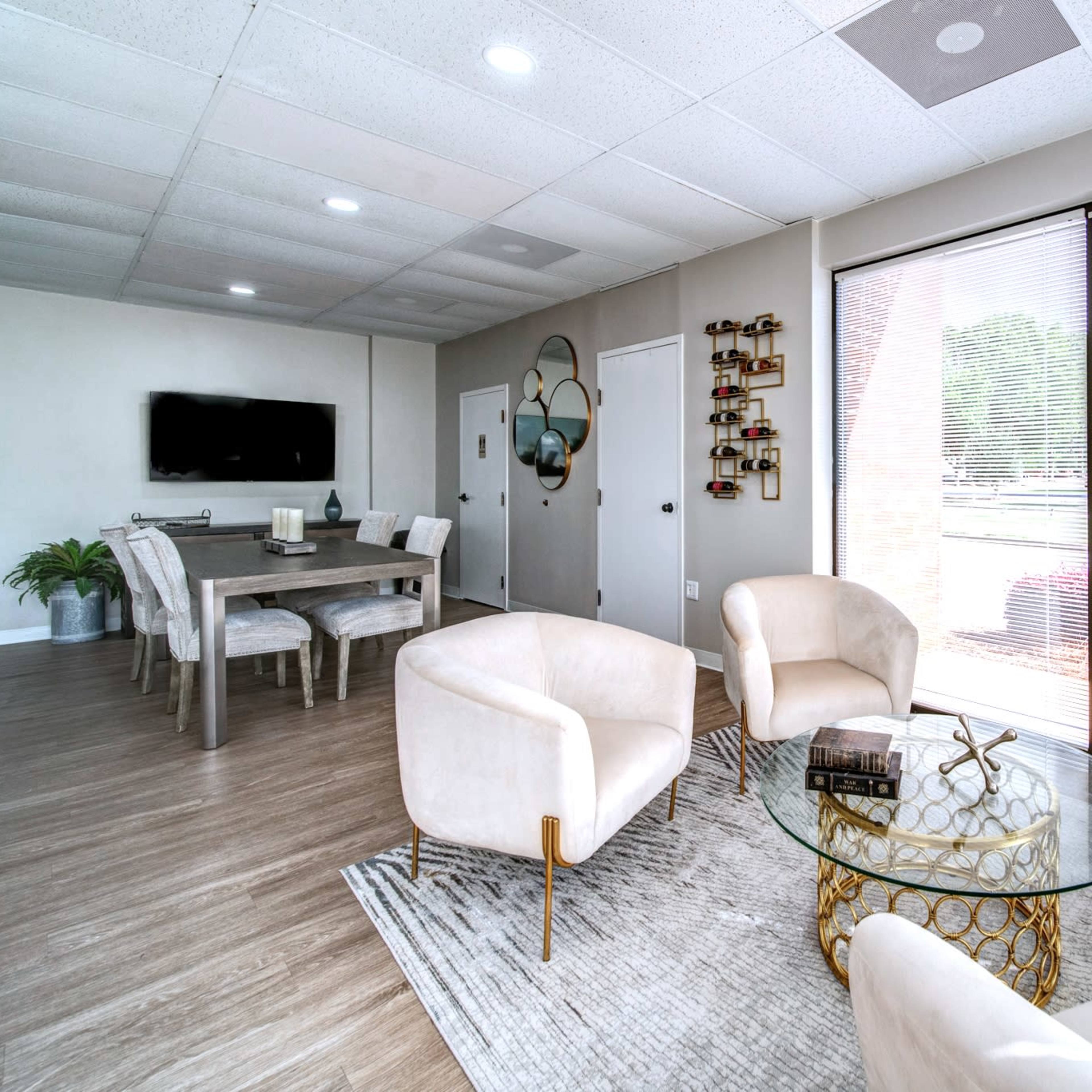 A modern living area featuring a round coffee table, four upholstered chairs, a dining table in the background, and large windows with blinds.