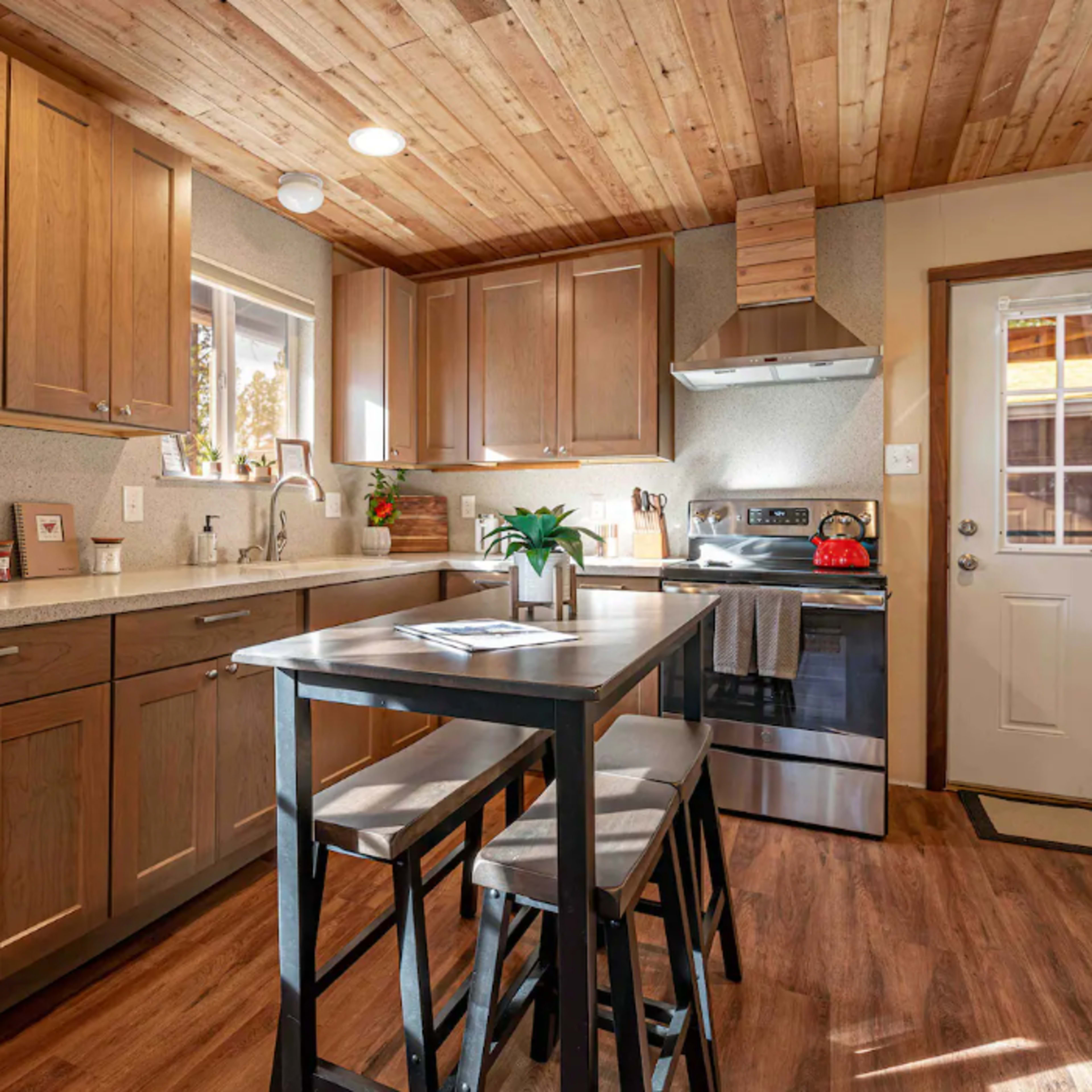 A modern kitchen features wooden cabinets, a stainless steel stove, and a small dining table with stools, all illuminated by natural light from a window.