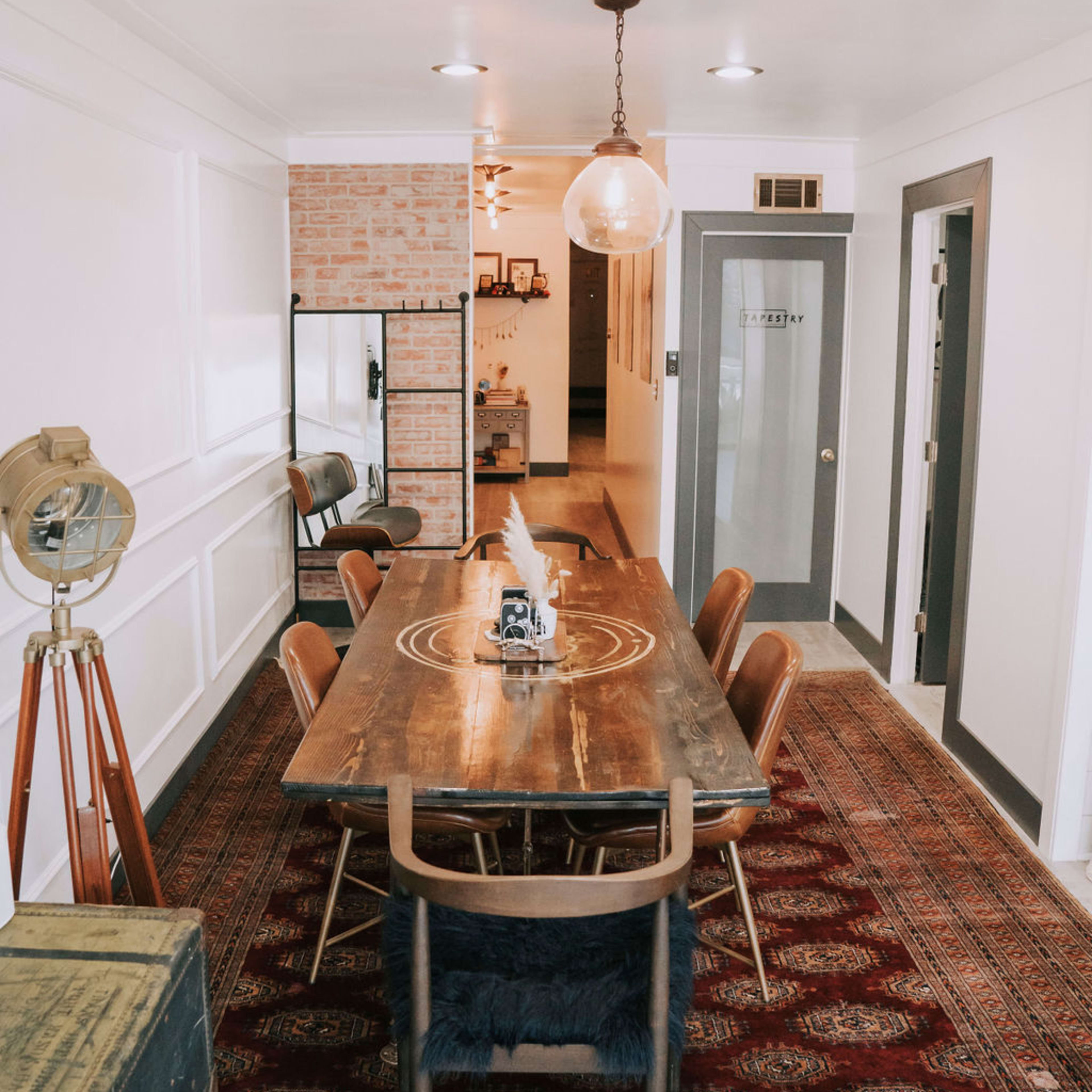 A dining area with a large wooden table surrounded by six chairs, set against a background of brick and white walls, with a rug on the floor and a hanging light fixture above.