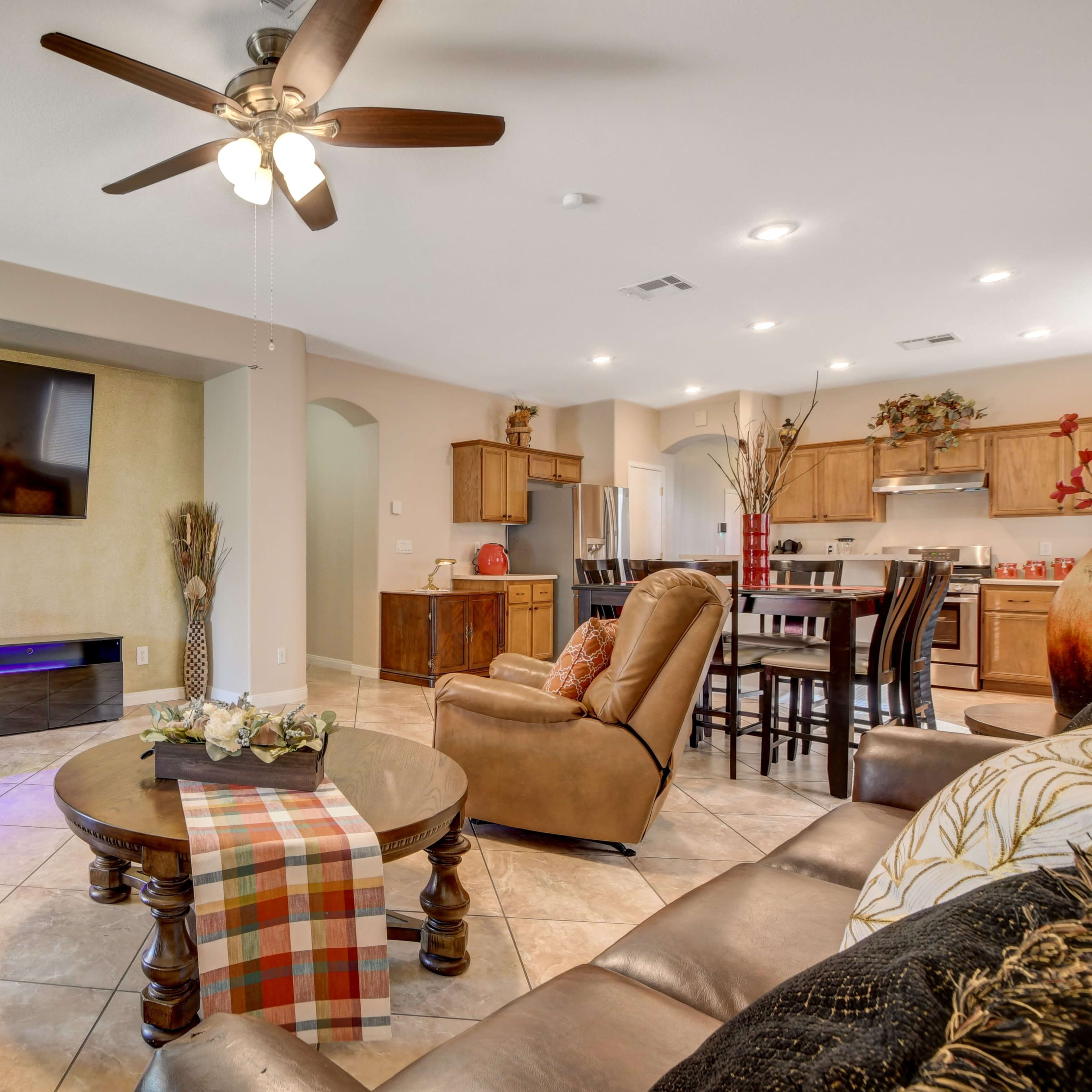The scene shows a living room featuring a seating area with two armchairs and a coffee table, a wall-mounted television, and an open kitchen in the background.