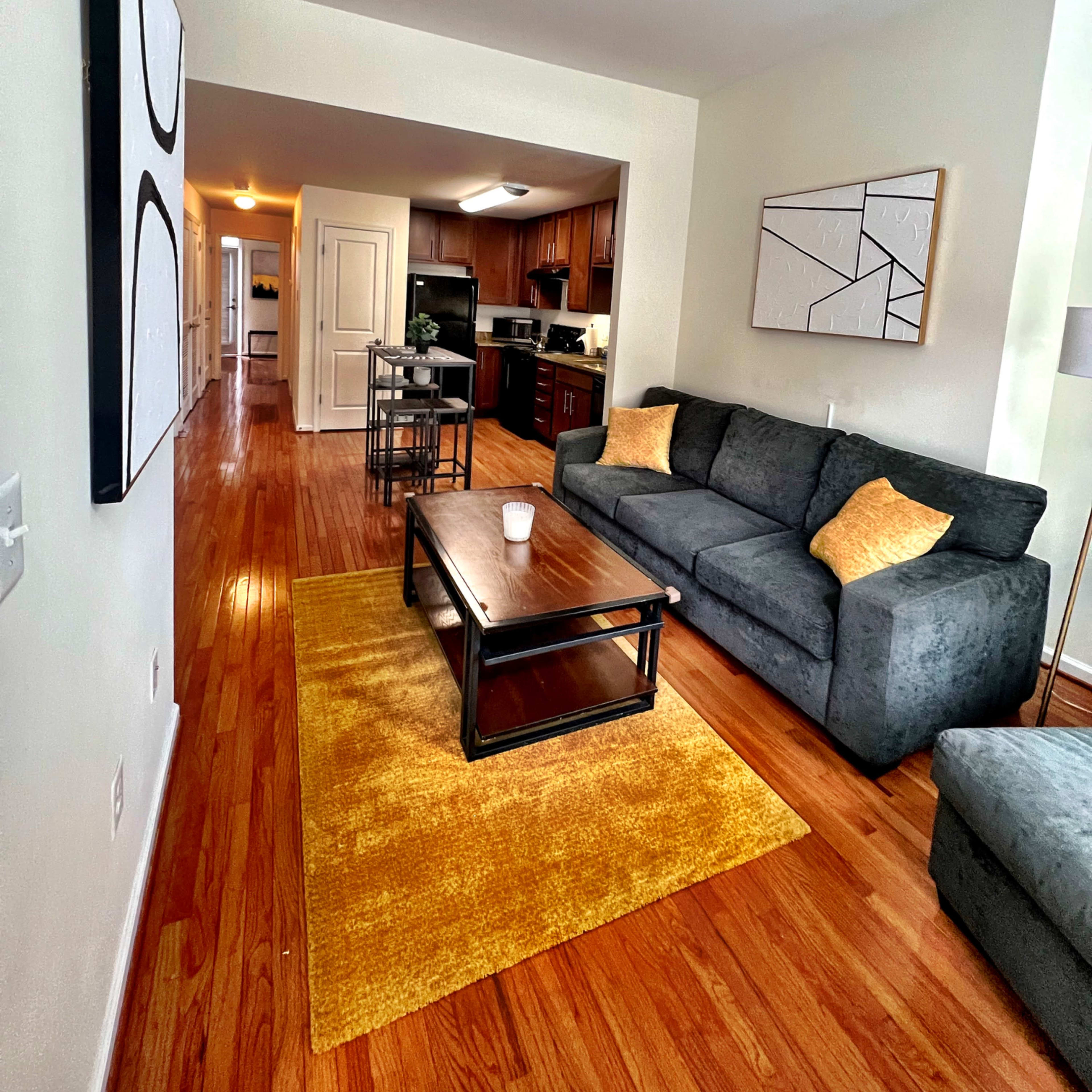 A living room with two gray sofas, a coffee table, a yellow rug, and an open kitchen in the background.