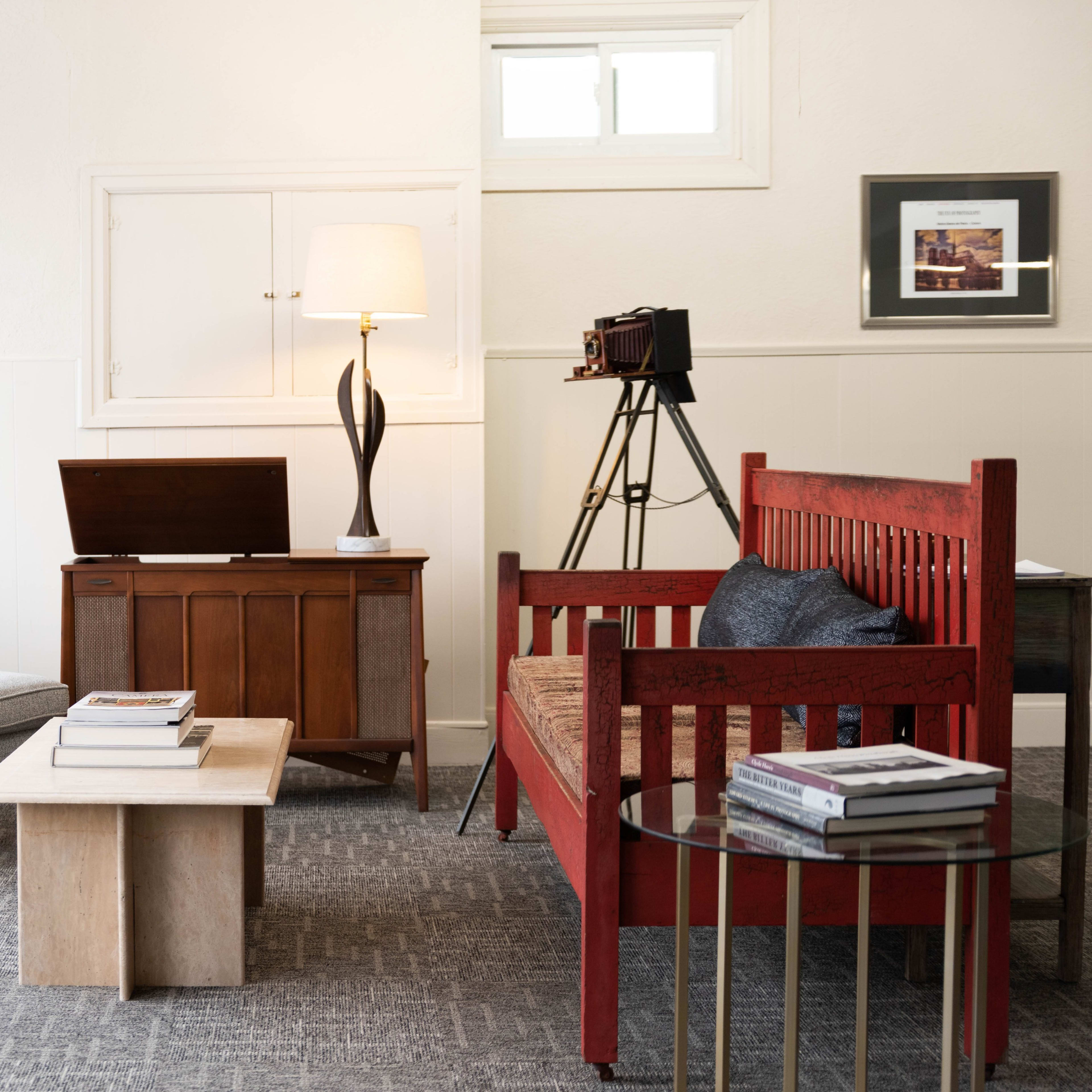 A modern living space featuring a mix of vintage and contemporary furniture, including a red wooden bench, a gray armchair, a wooden cabinet, and a tripod camera.