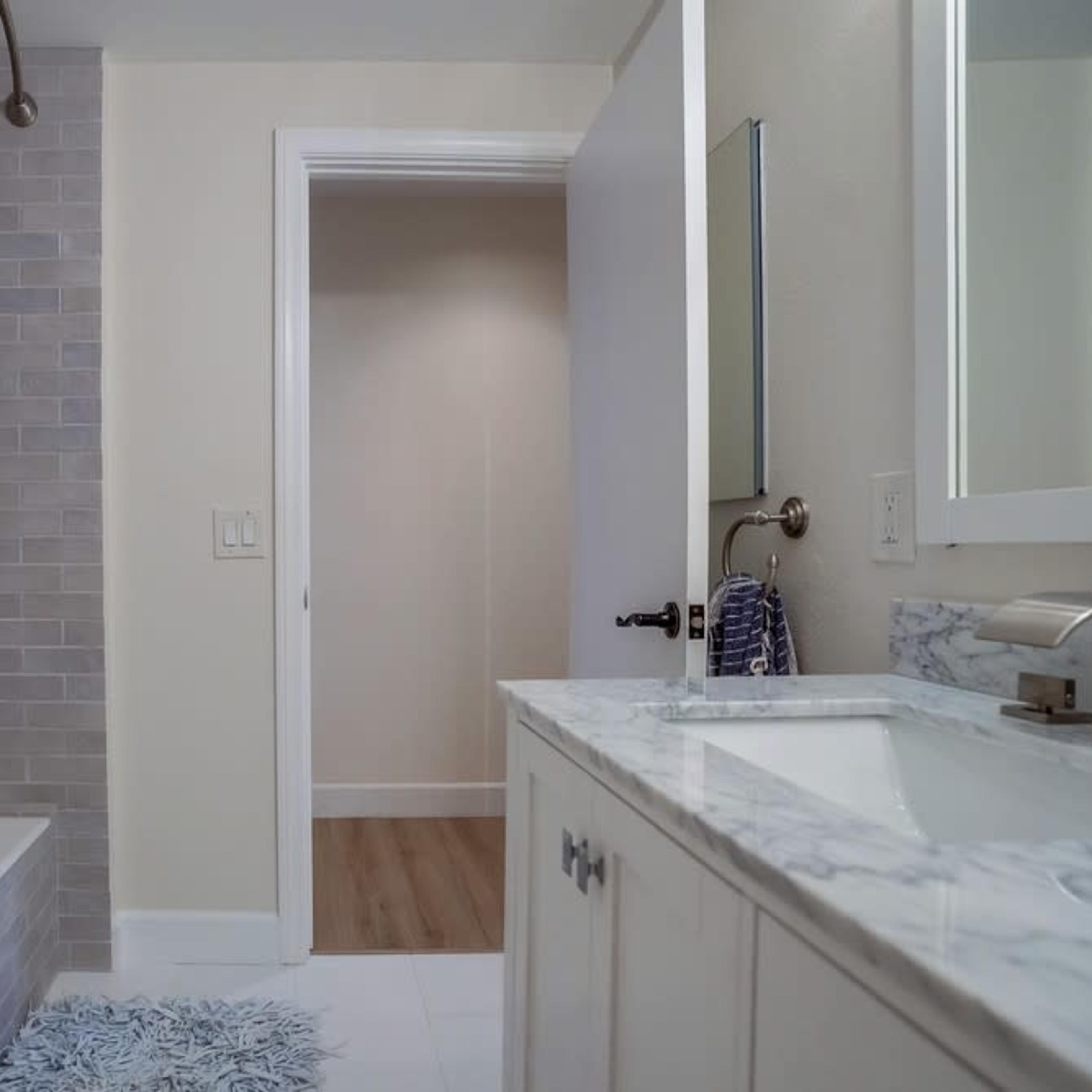 A bathroom featuring a bathtub with a shower curtain, a marble countertop with a sink, and a doorway leading to another room.