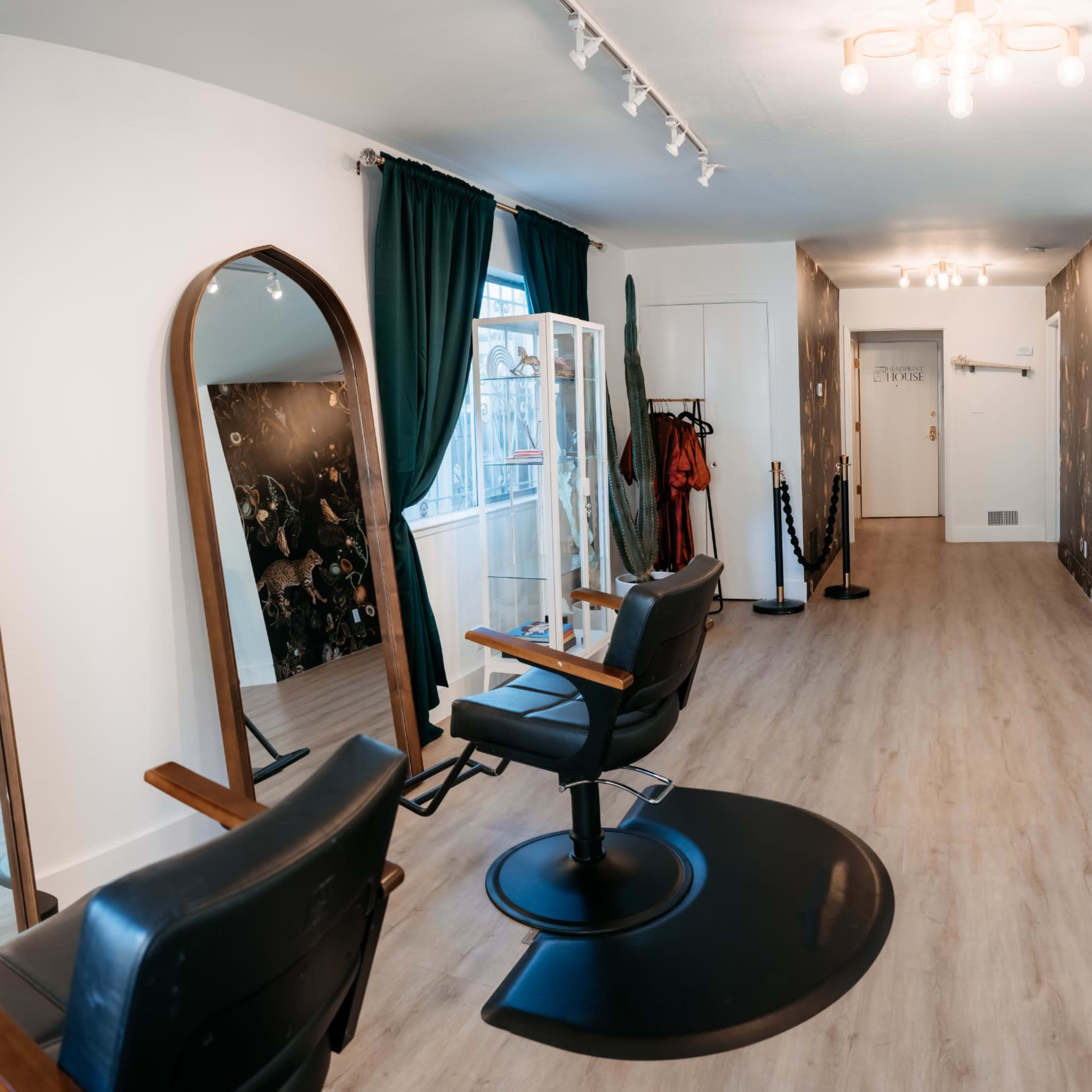 A spacious and well-lit salon interior featuring two black salon chairs, a mirrored wall, green curtains, and a brown leather bench with decorative plants.