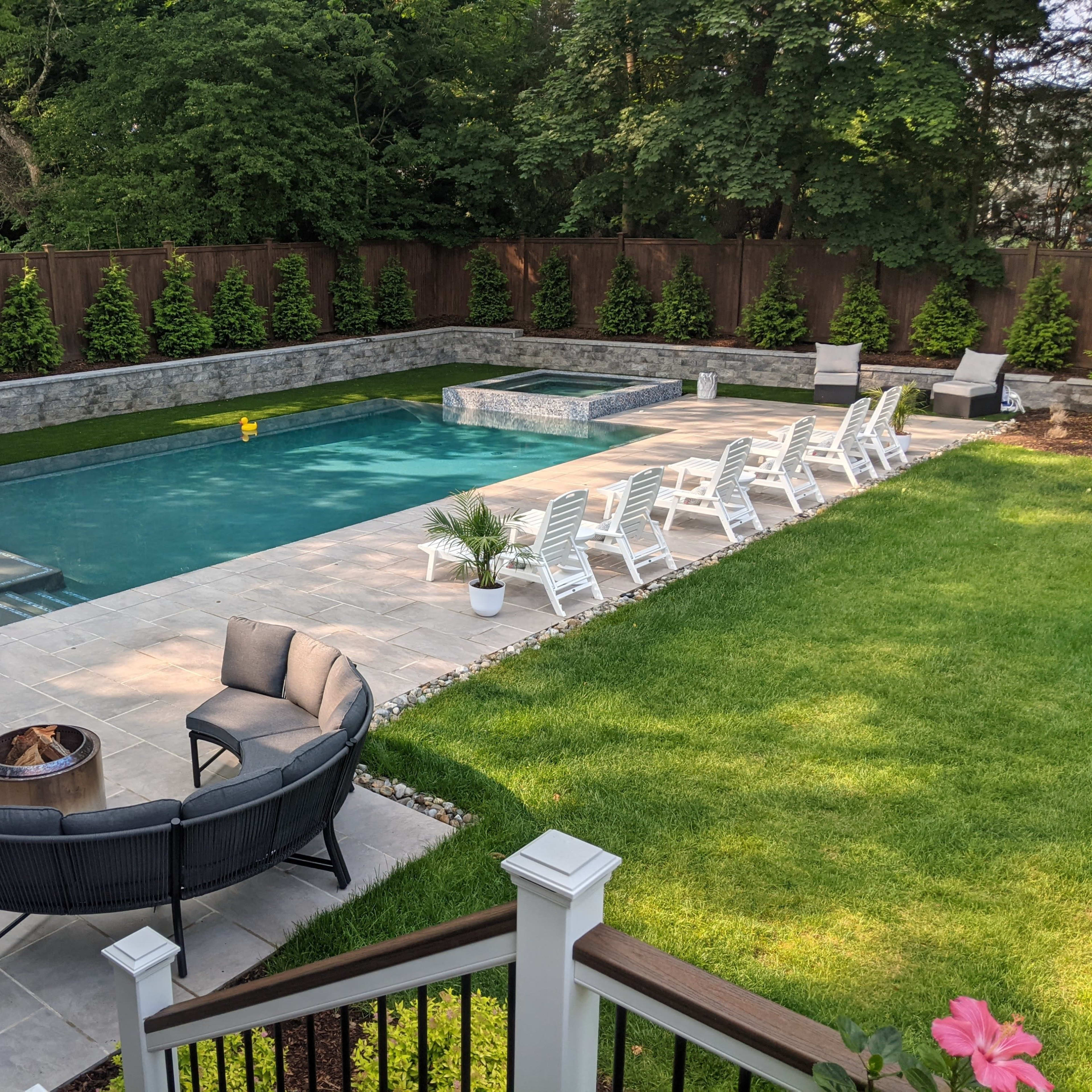 A backyard with a rectangular swimming pool surrounded by lounge chairs, stone paths, and landscaped greenery.