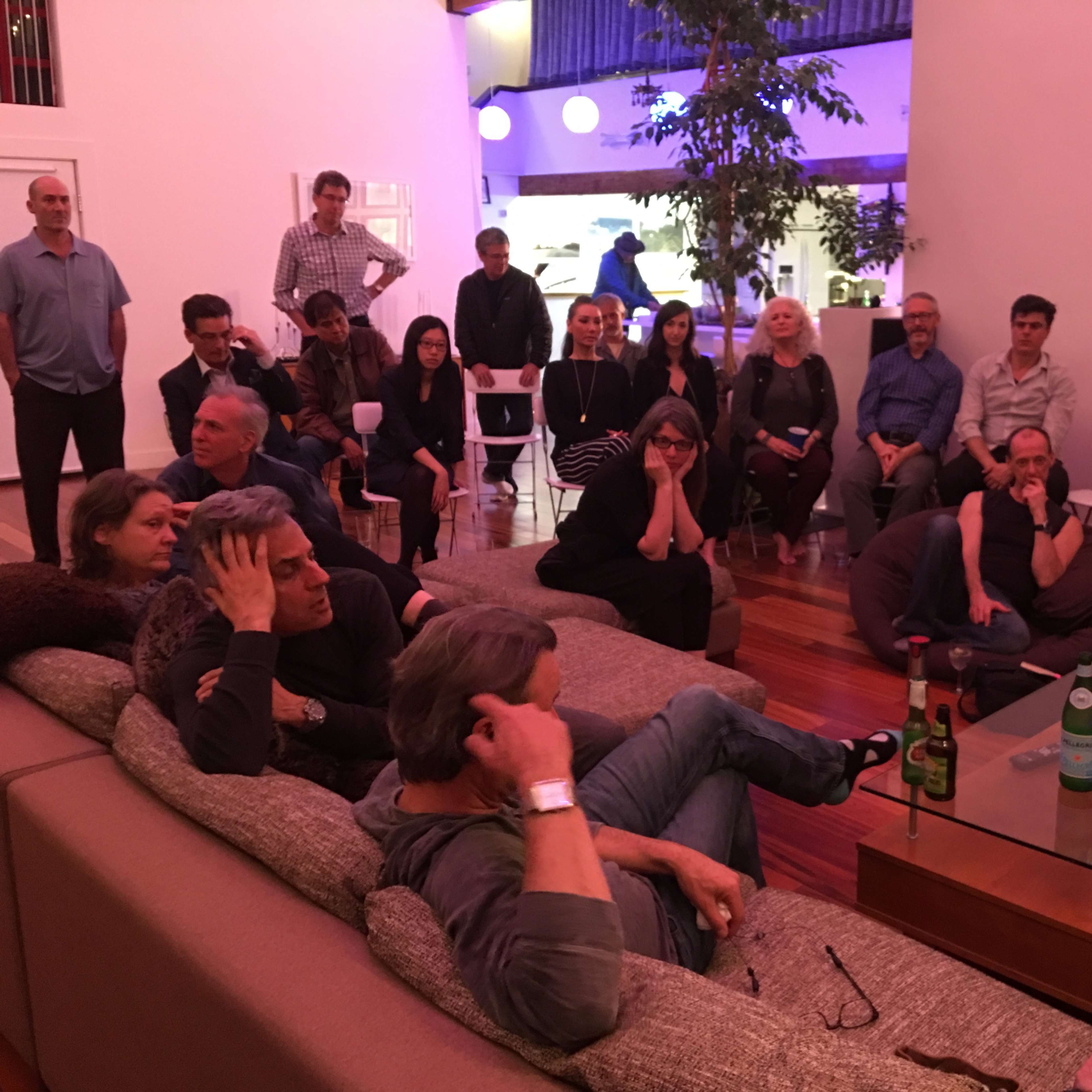 A group of people is gathered in a living room, some seated on sofas and bean bags while others are standing and facing the center of the room.