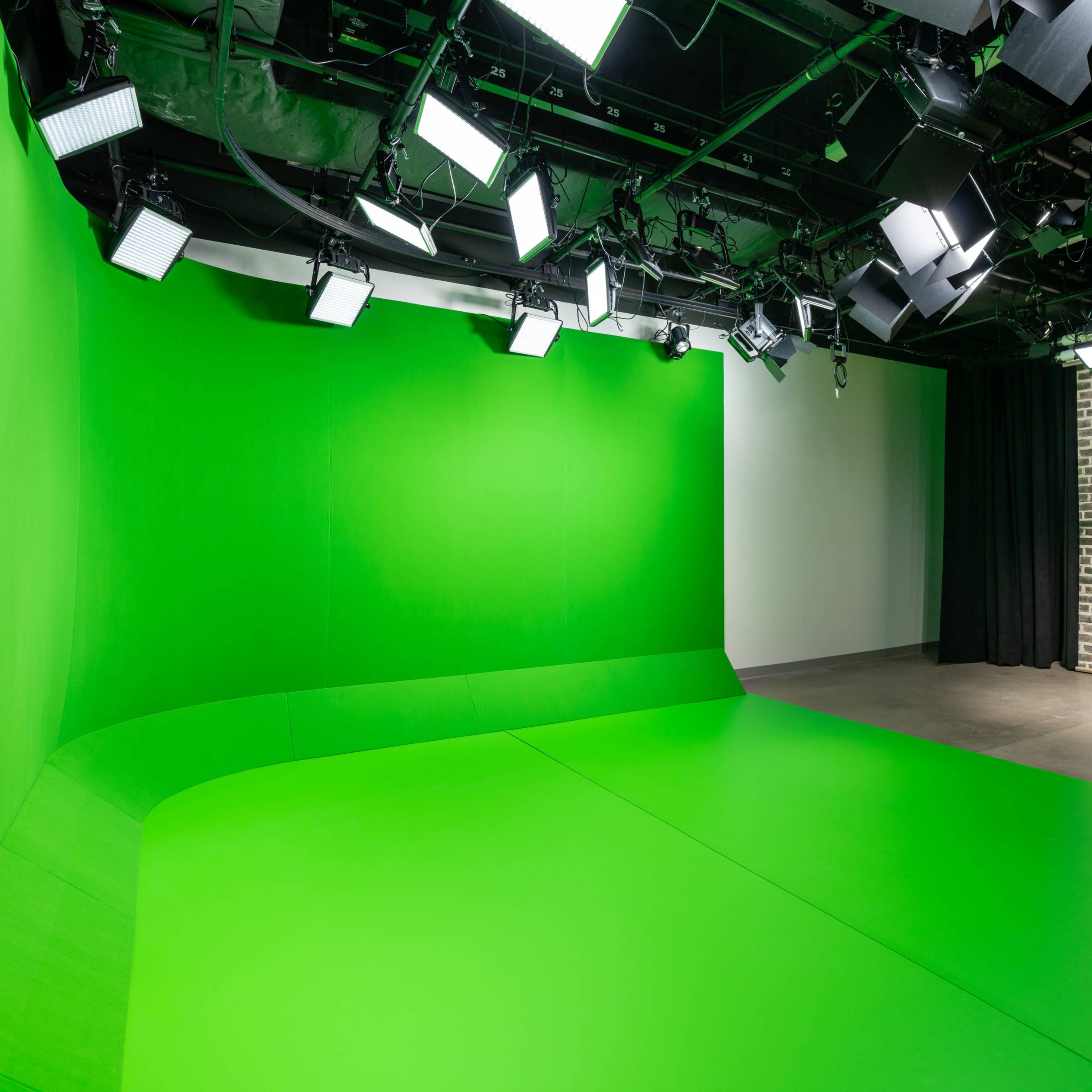 A studio featuring a green screen backdrop, multiple overhead lights, and a brick wall on one side.