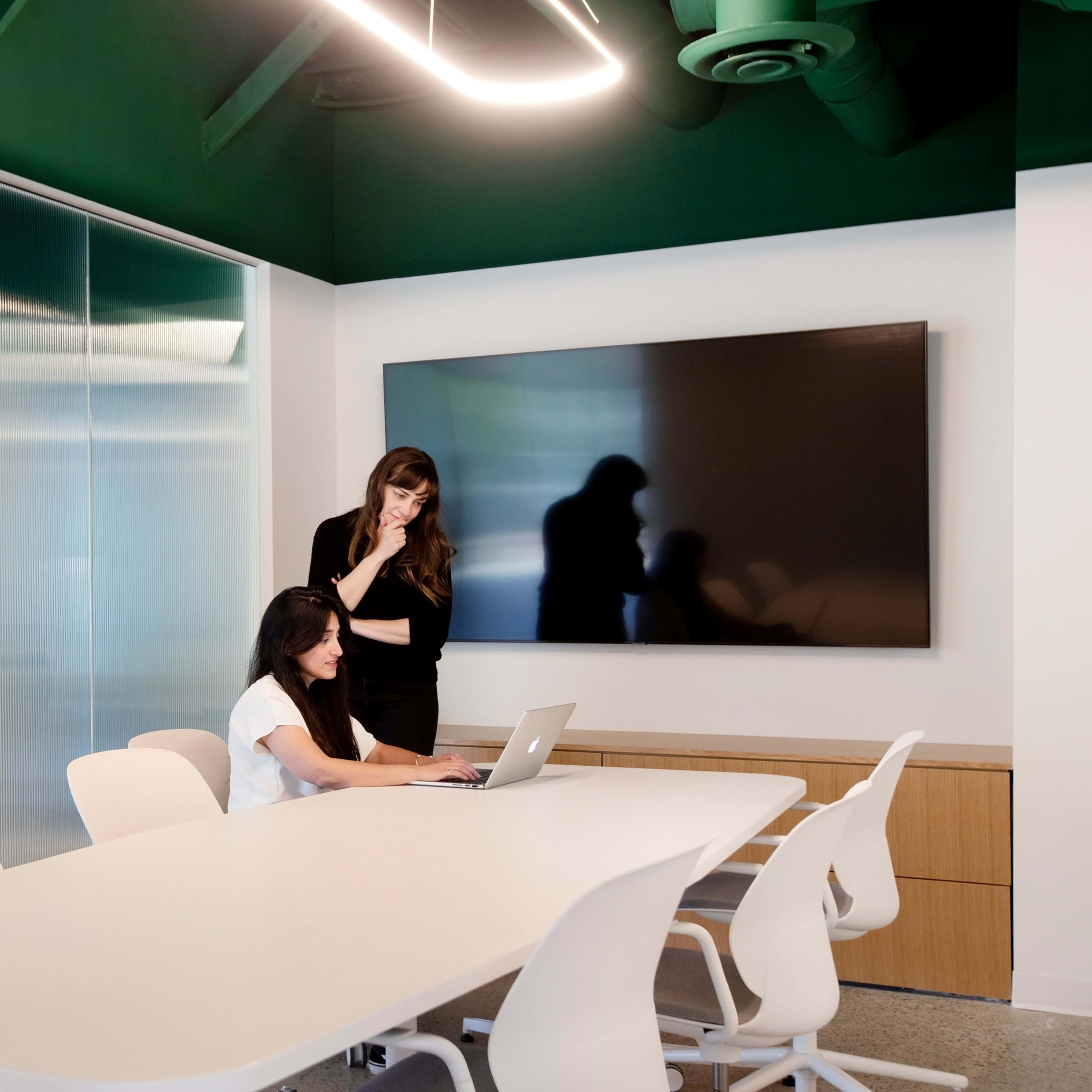 Two women are collaborating in a modern conference room with a large screen and a long table.