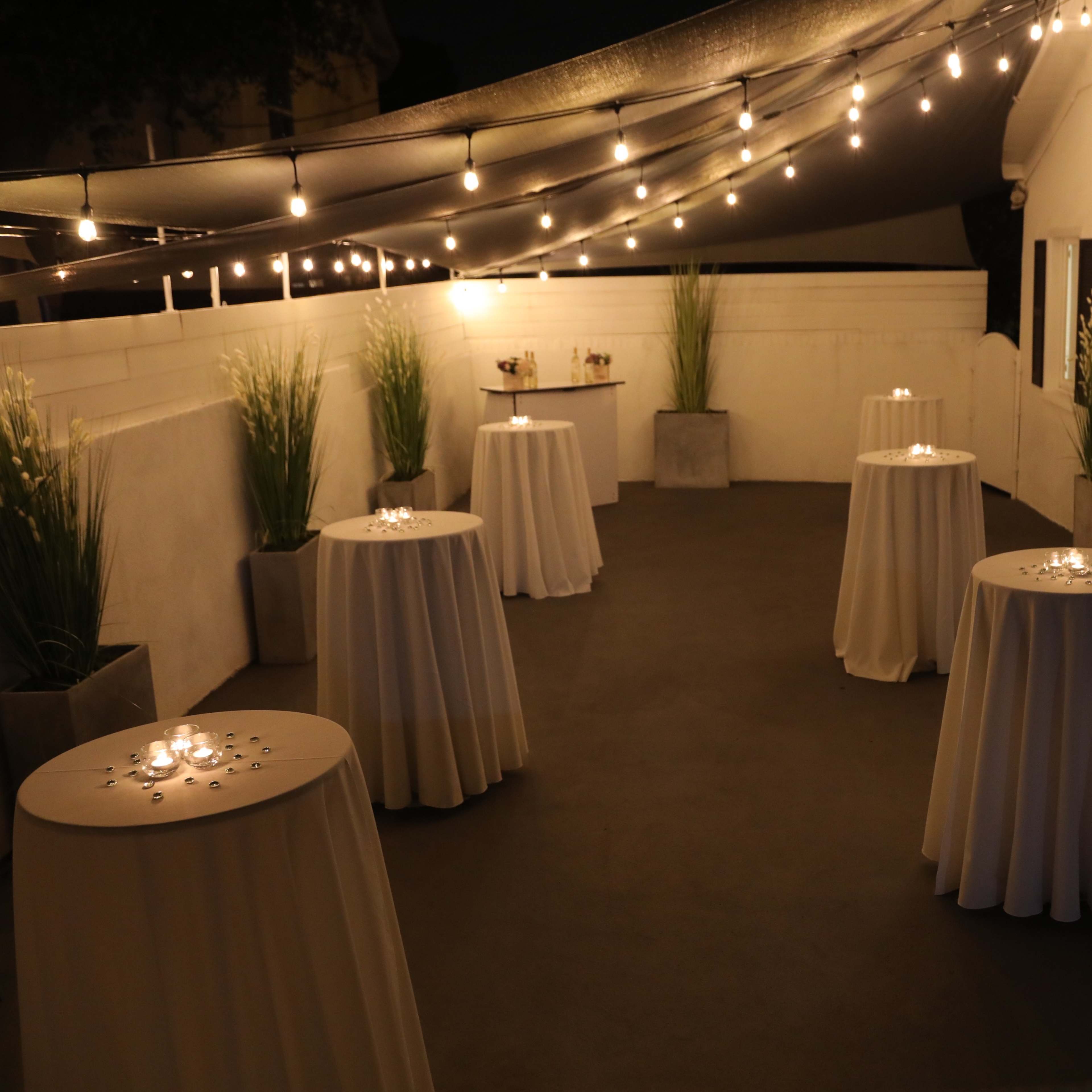 A dimly lit outdoor space with several round tables covered in white tablecloths, surrounded by planters and string lights overhead.