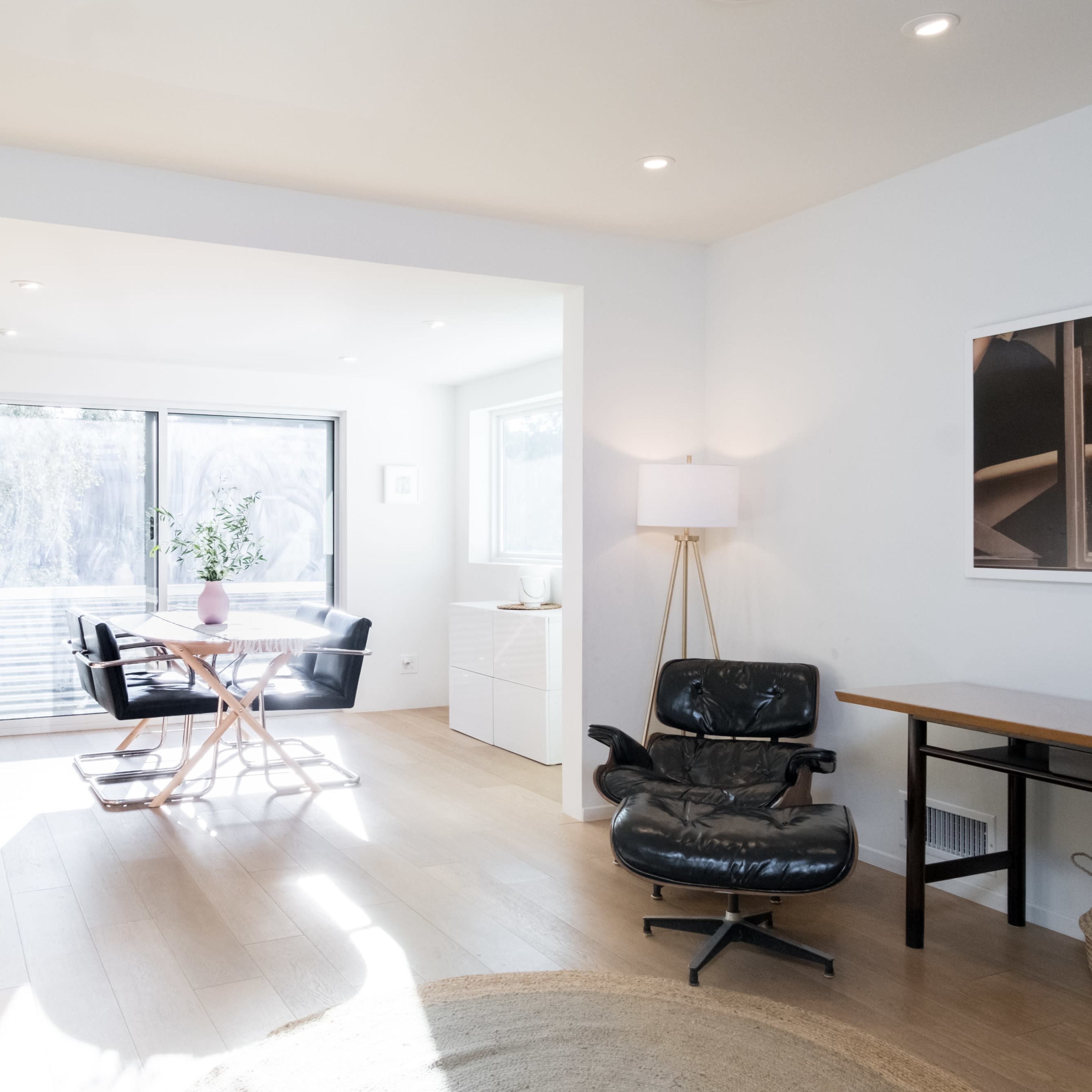 A modern, open living space with a light-colored sofa, a black chair, a circular rug, and a dining area featuring a round table and chairs, illuminated by natural light from large windows.
