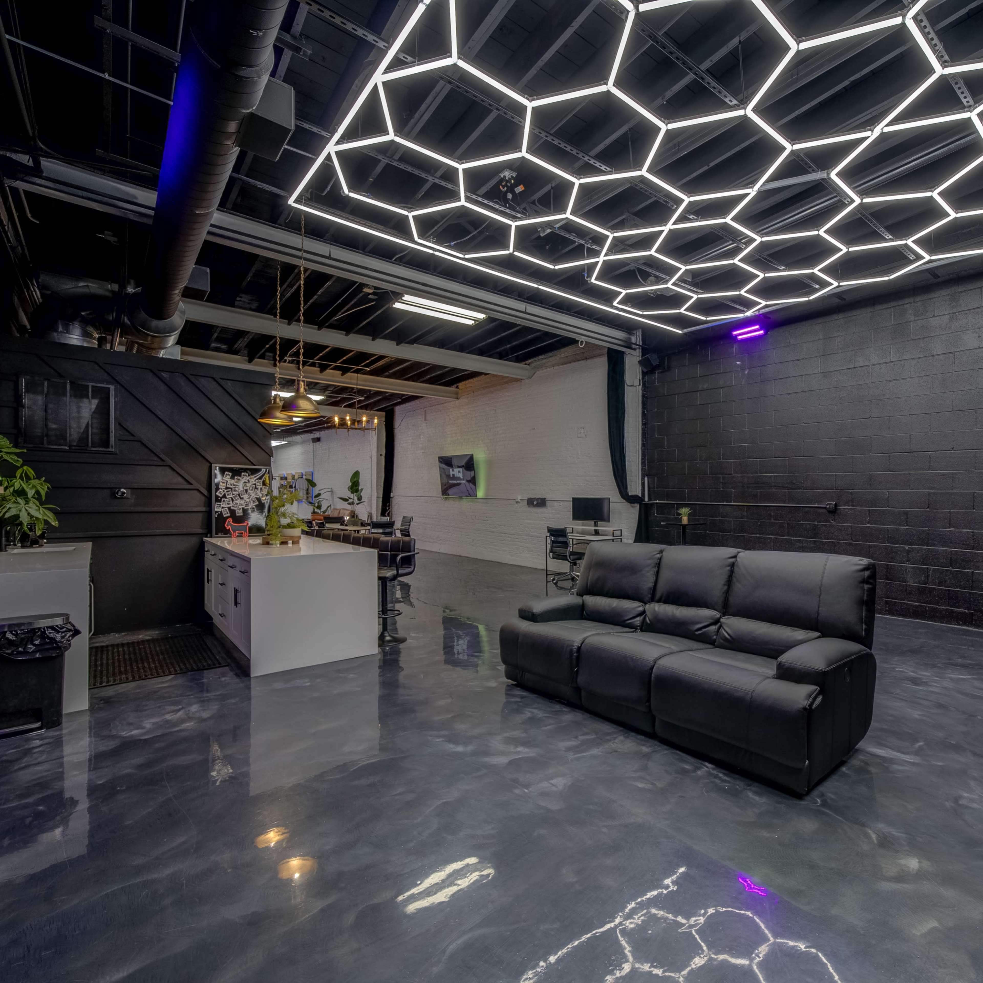 A modern lounge area features a black reclining sofa beneath a hexagonal lighted ceiling in an industrial-style room with a polished concrete floor.