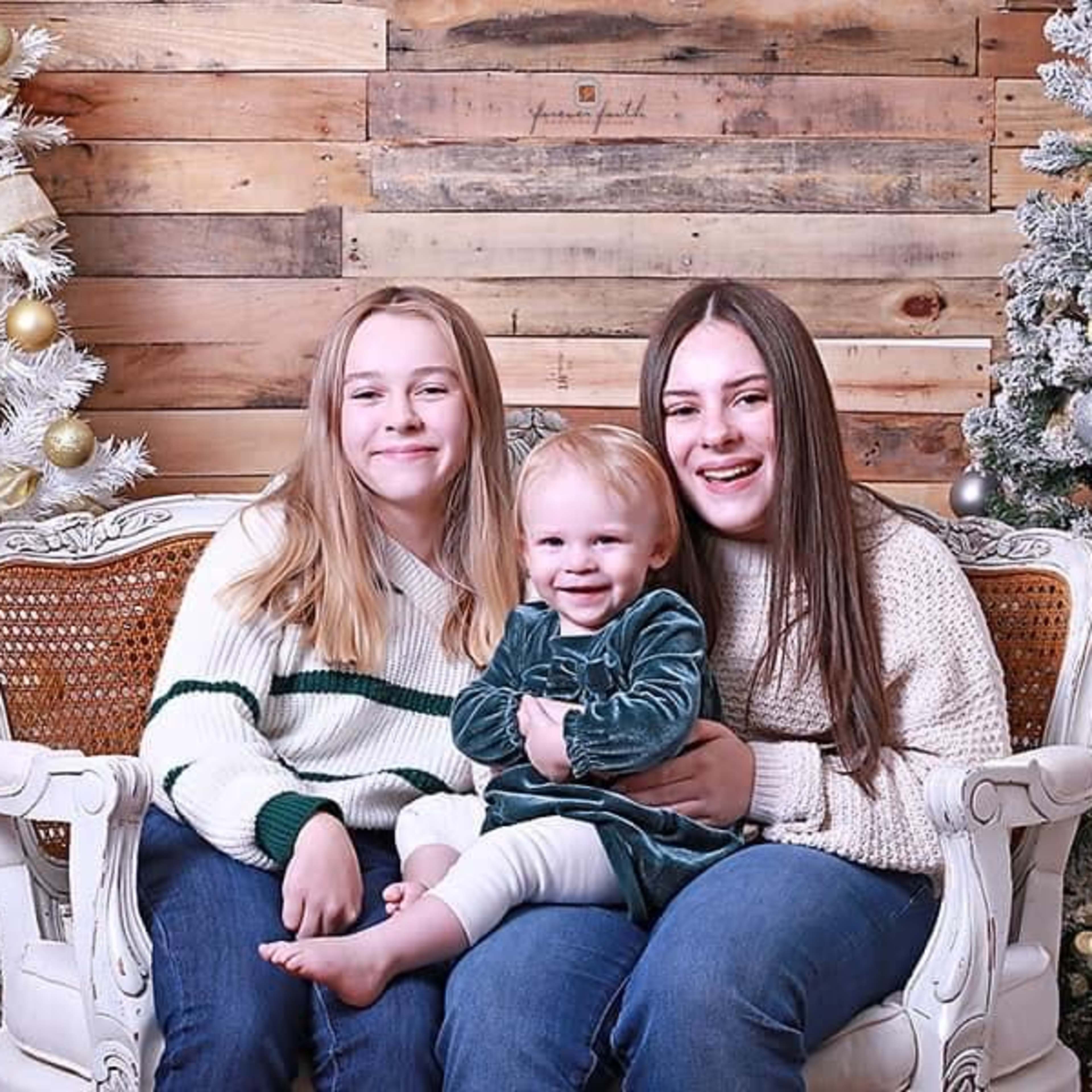 Two girls sit on a sofa beside a toddler, with decorated Christmas trees in the background.