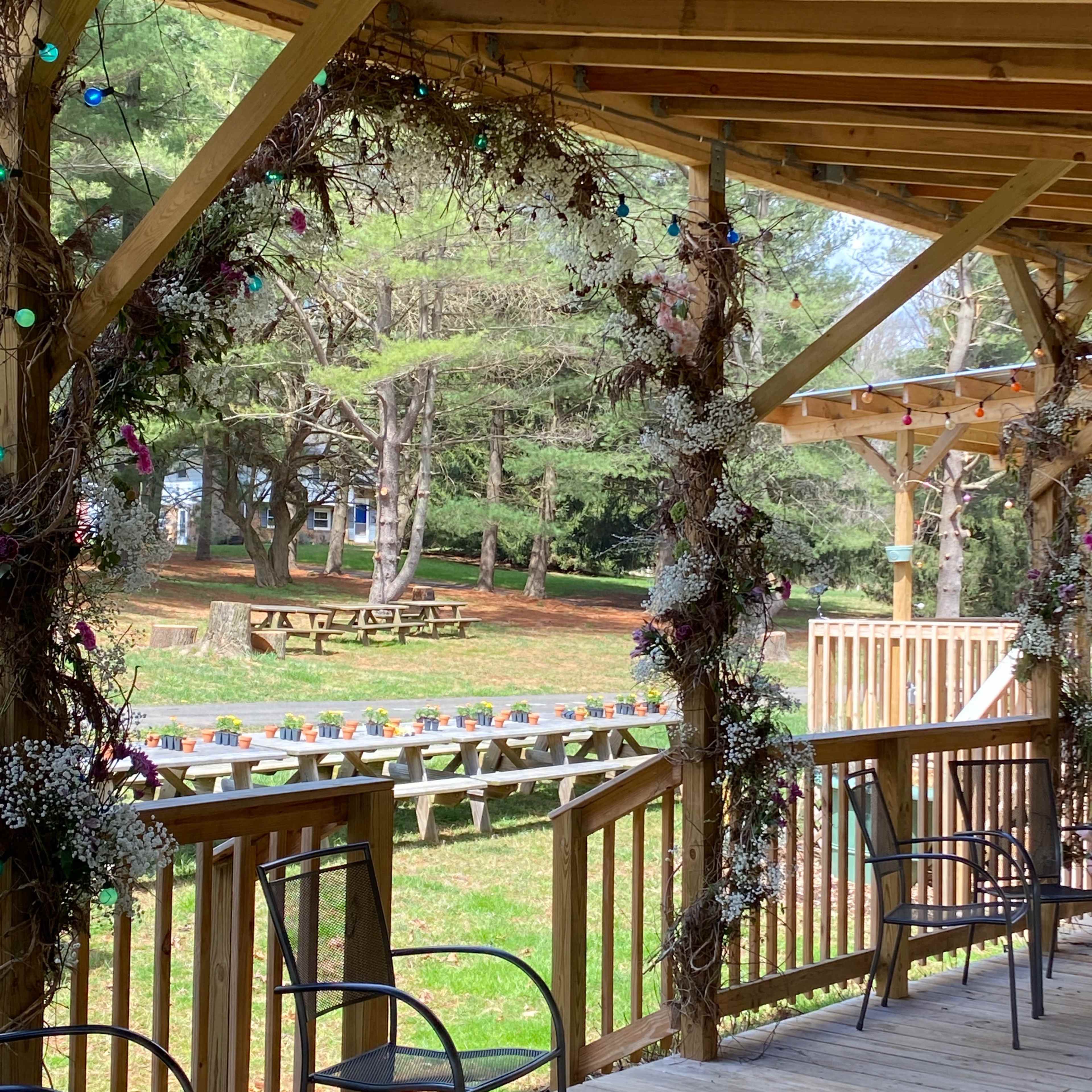 A wooden porch with black chairs overlooks a grassy area with picnic tables and flower planters in the background.