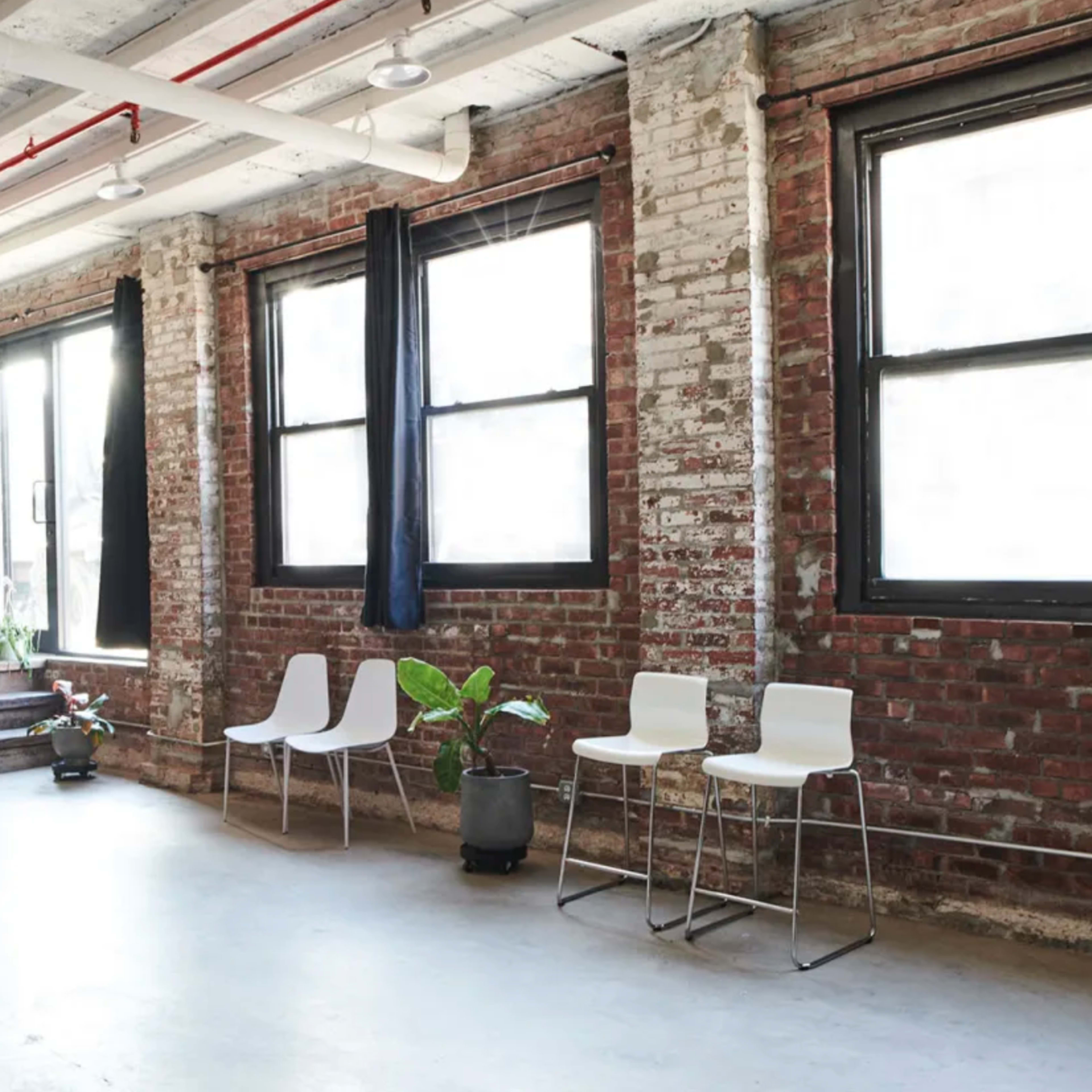 A room with exposed brick walls, large windows, and three white chairs positioned near a staircase and potted plants.
