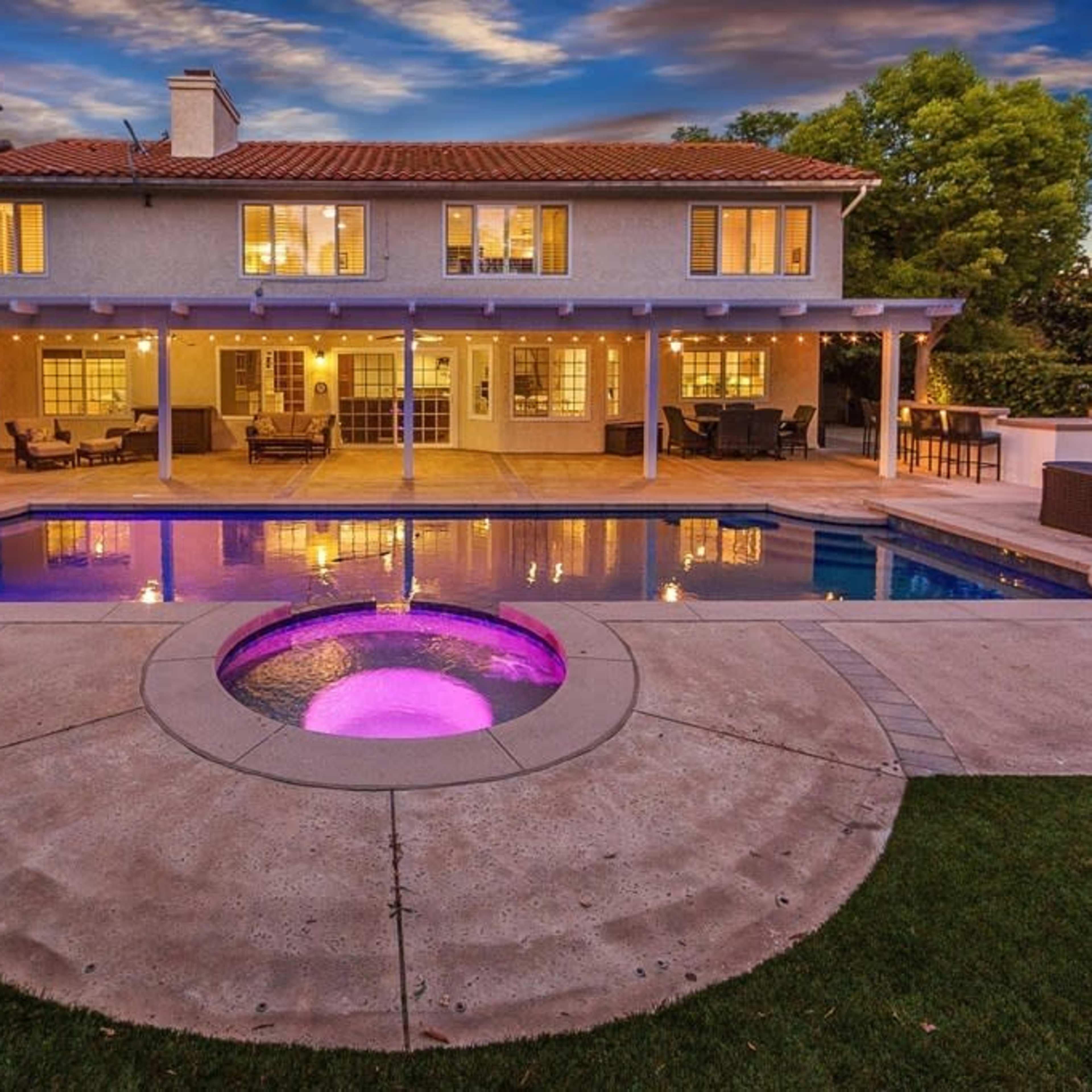 A two-story house with a tiled roof overlooks a backyard featuring a swimming pool and a hot tub surrounded by landscaped greenery.
