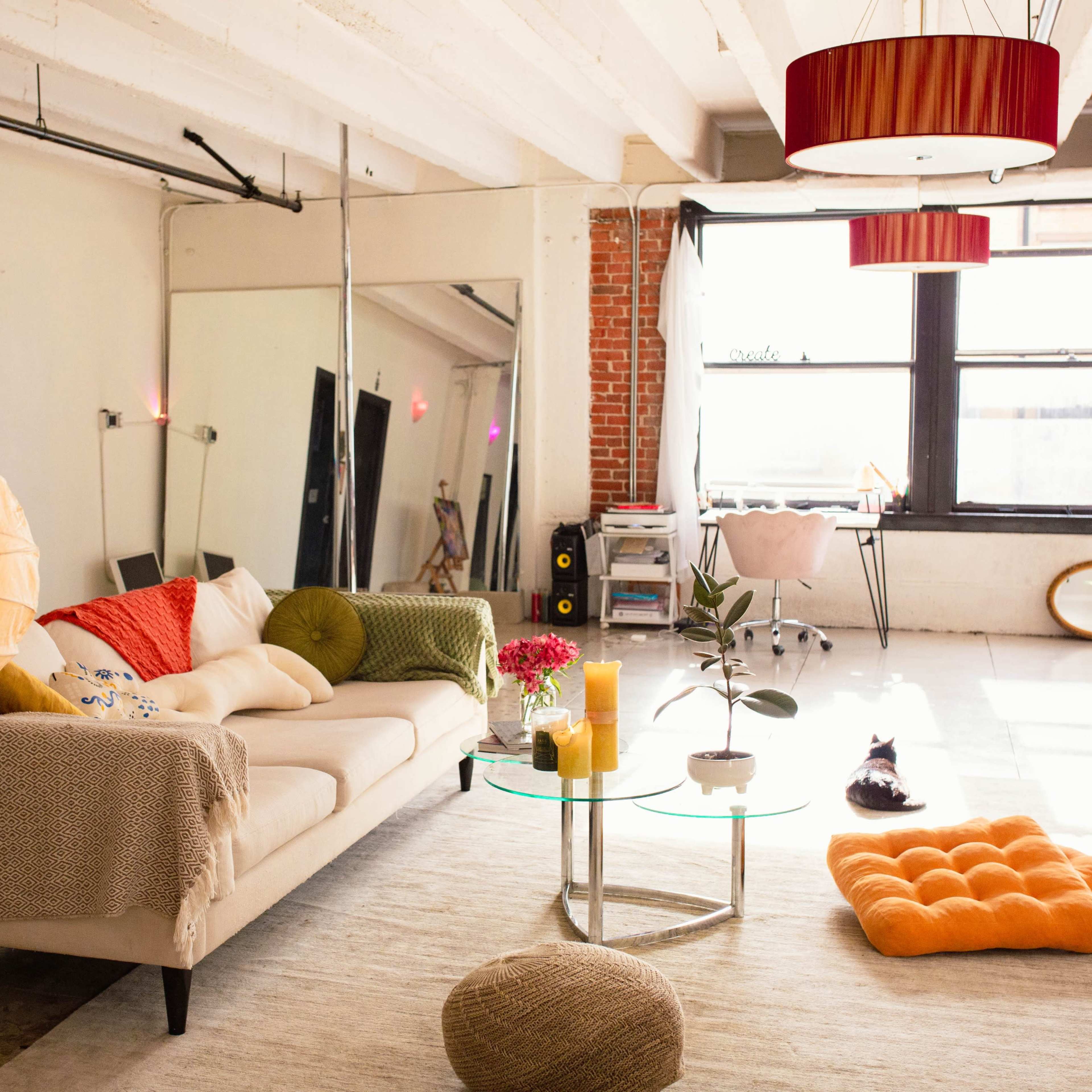 A bright, open-plan living room featuring a sofa, a glass coffee table, colorful cushions, and large windows with natural light.