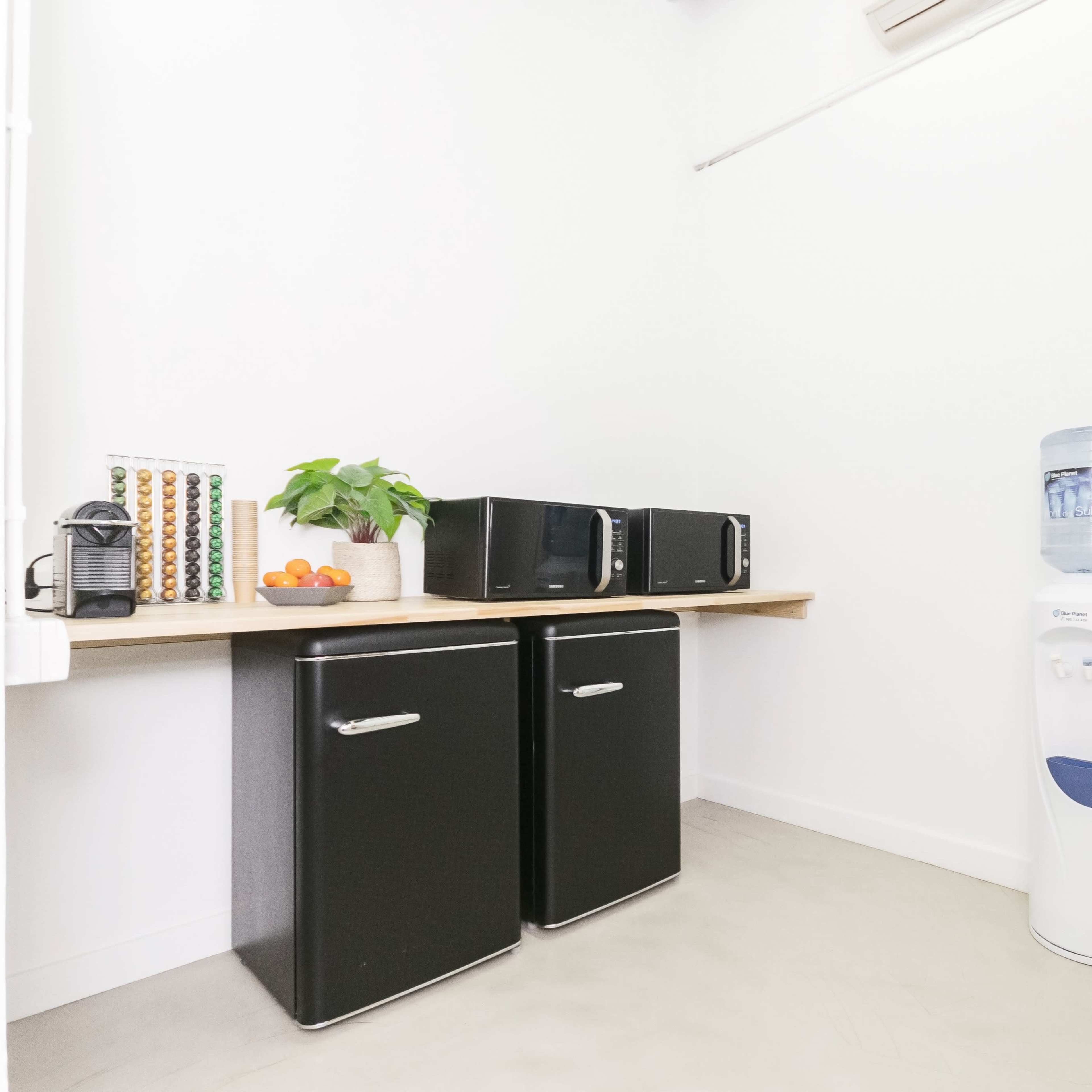 A small room features two black refrigerators, a countertop with a coffee machine and oranges, and a water cooler.