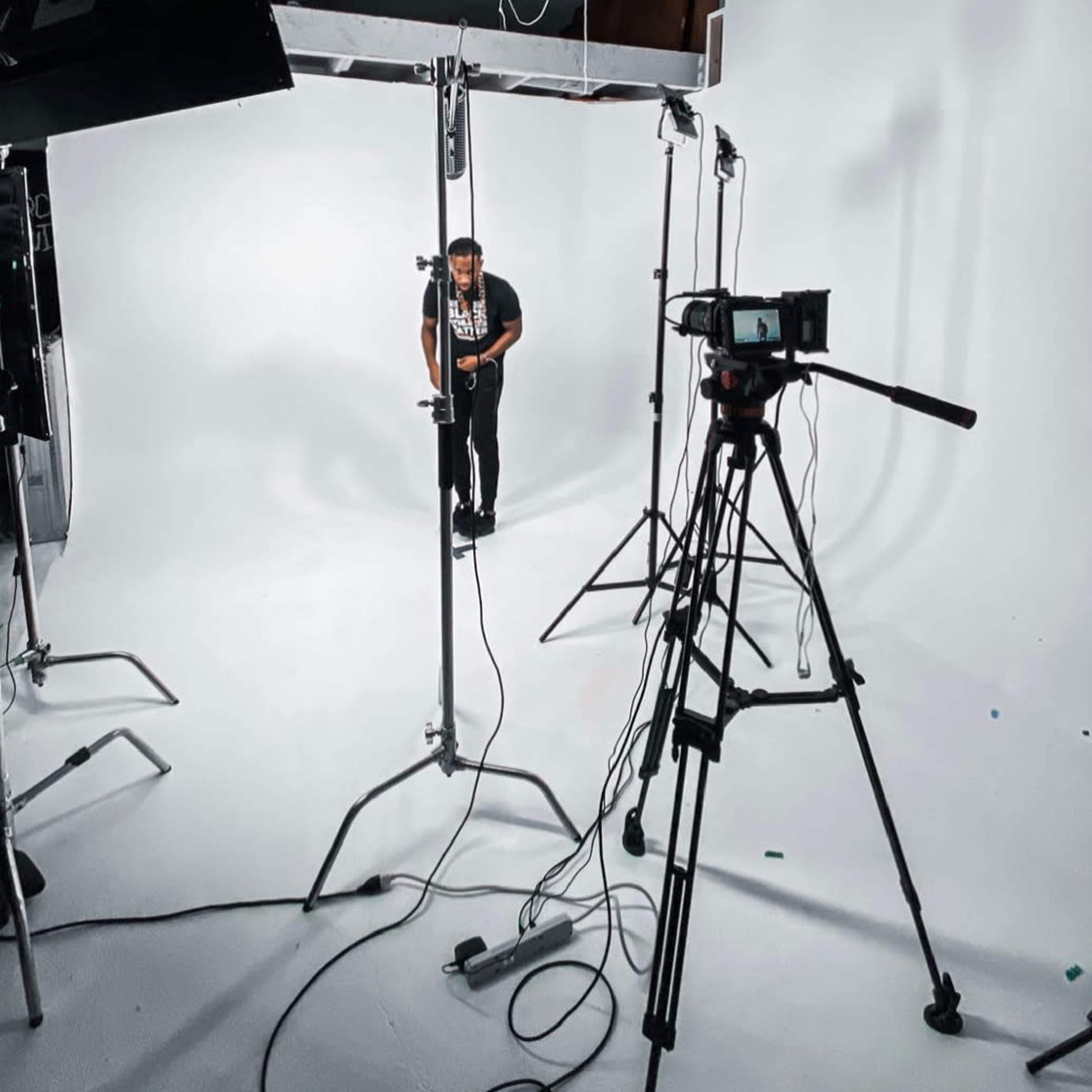 A person stands in a studio setting surrounded by lighting equipment and a camera on a tripod.