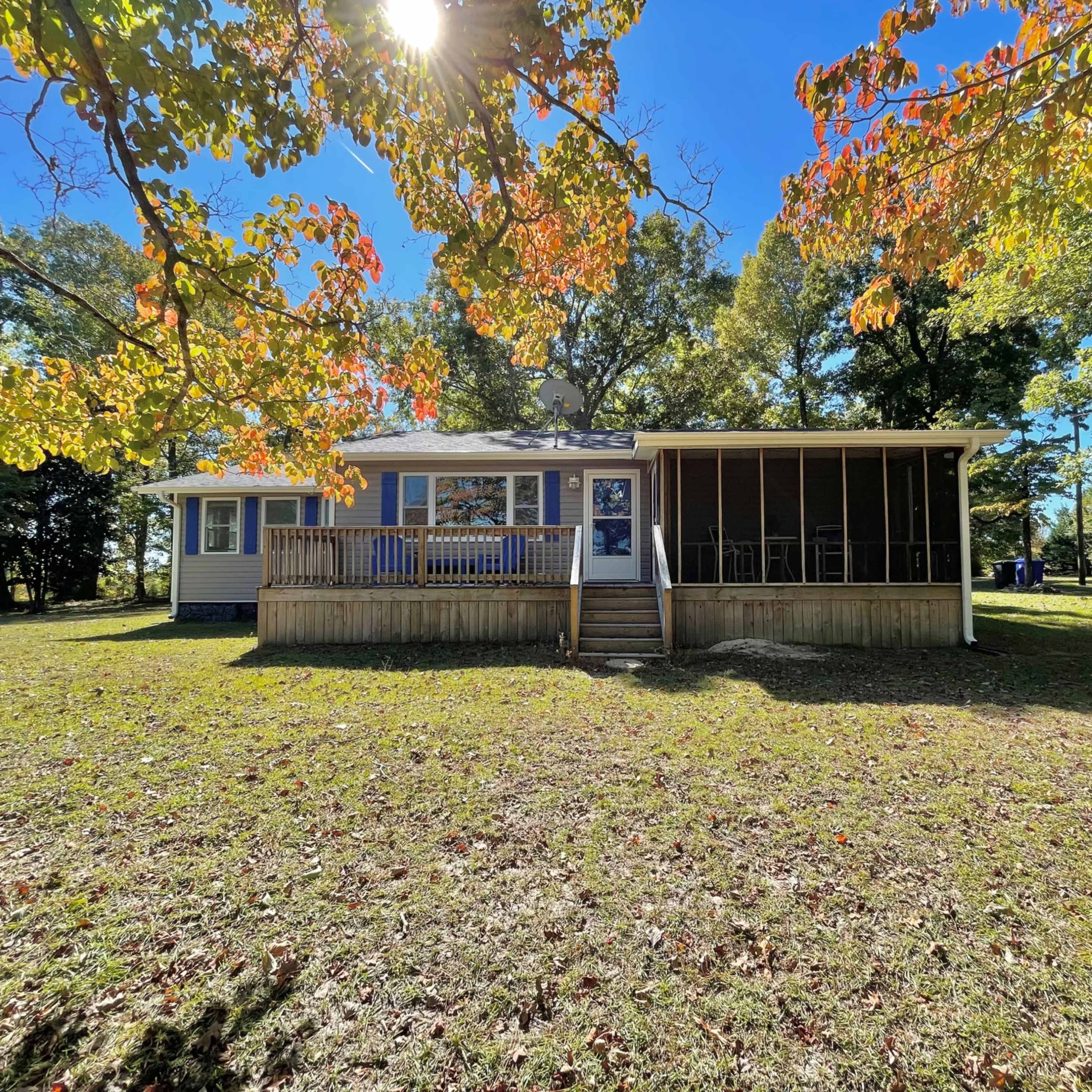 A single-story house with a screened porch and a front deck is surrounded by grass and trees.