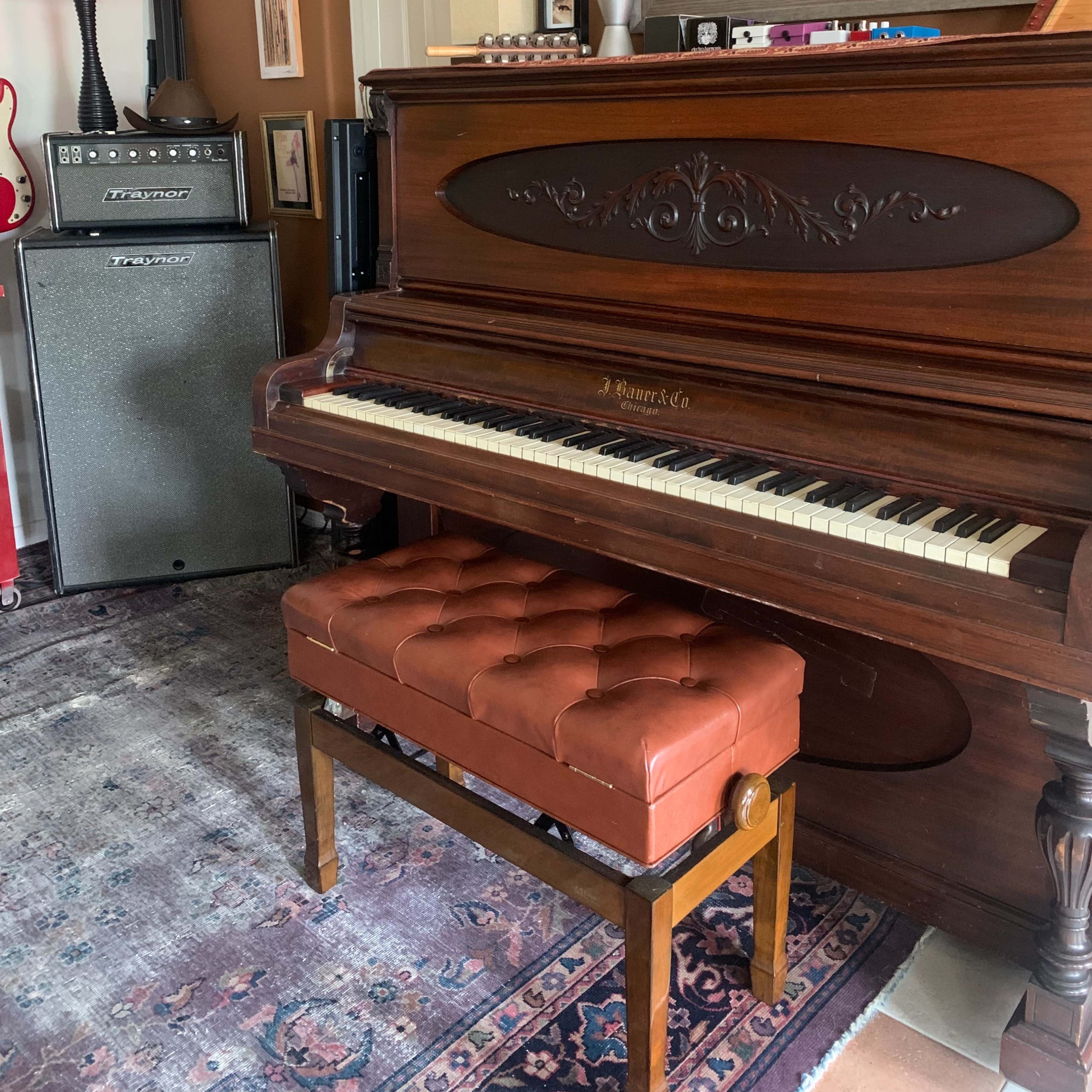A wooden piano with a tufted leather bench in front of it, situated next to an amplifier and a decorative mirror.
