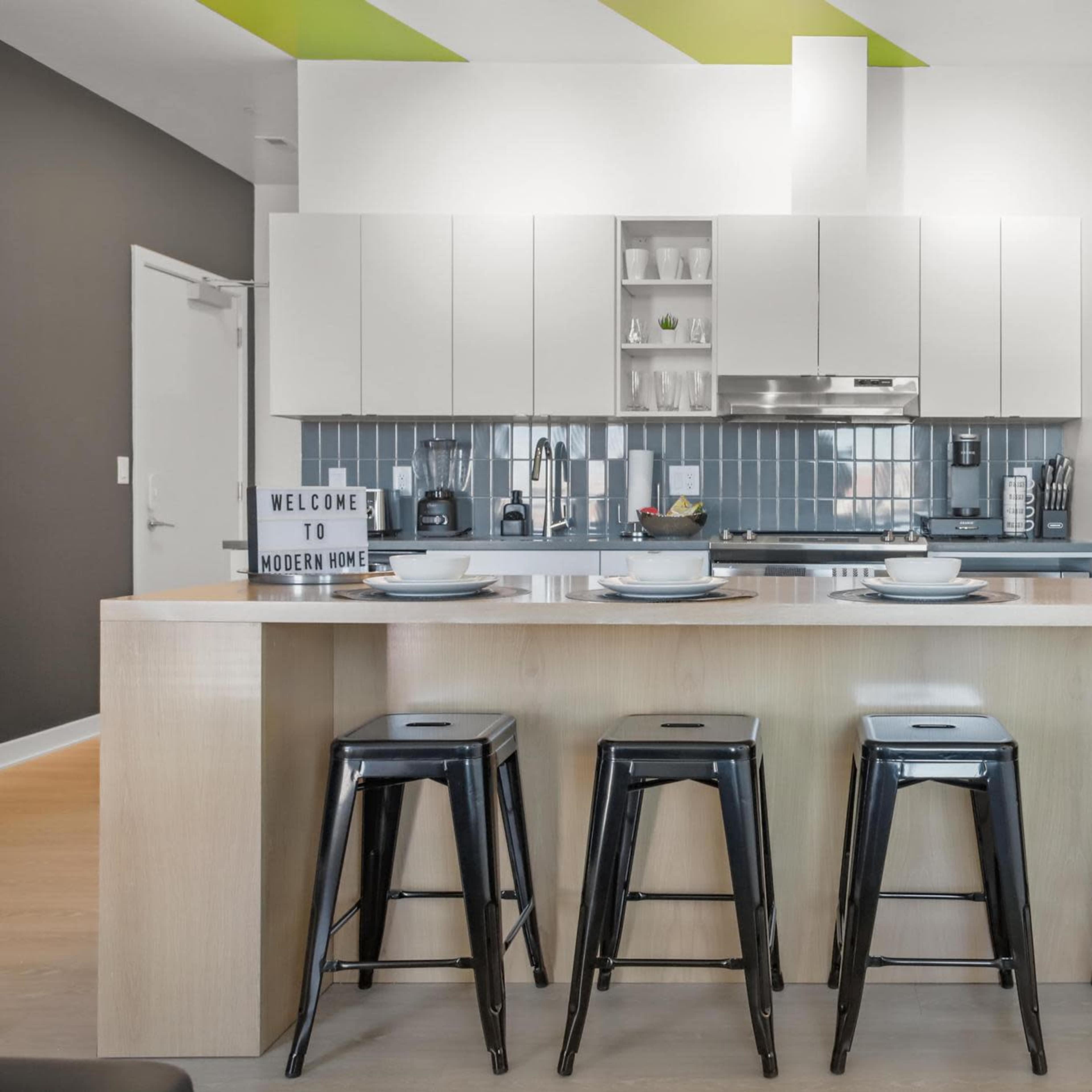 A modern kitchen features a central island with four black stools, white cabinets, and a stainless steel refrigerator.