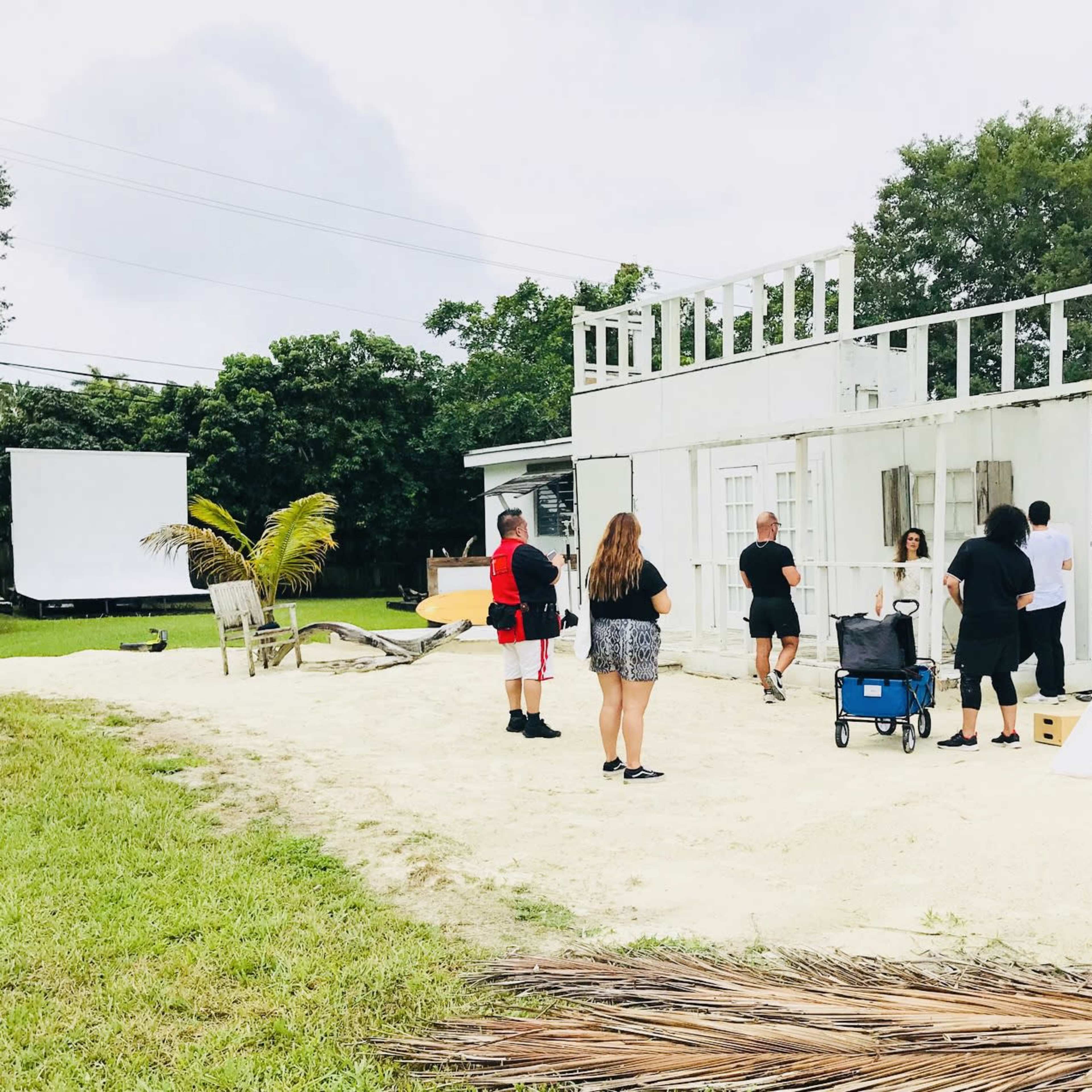 A group of people is preparing a photoshoot in an outdoor area featuring a partially constructed building, a teepee, and various props on the sand.