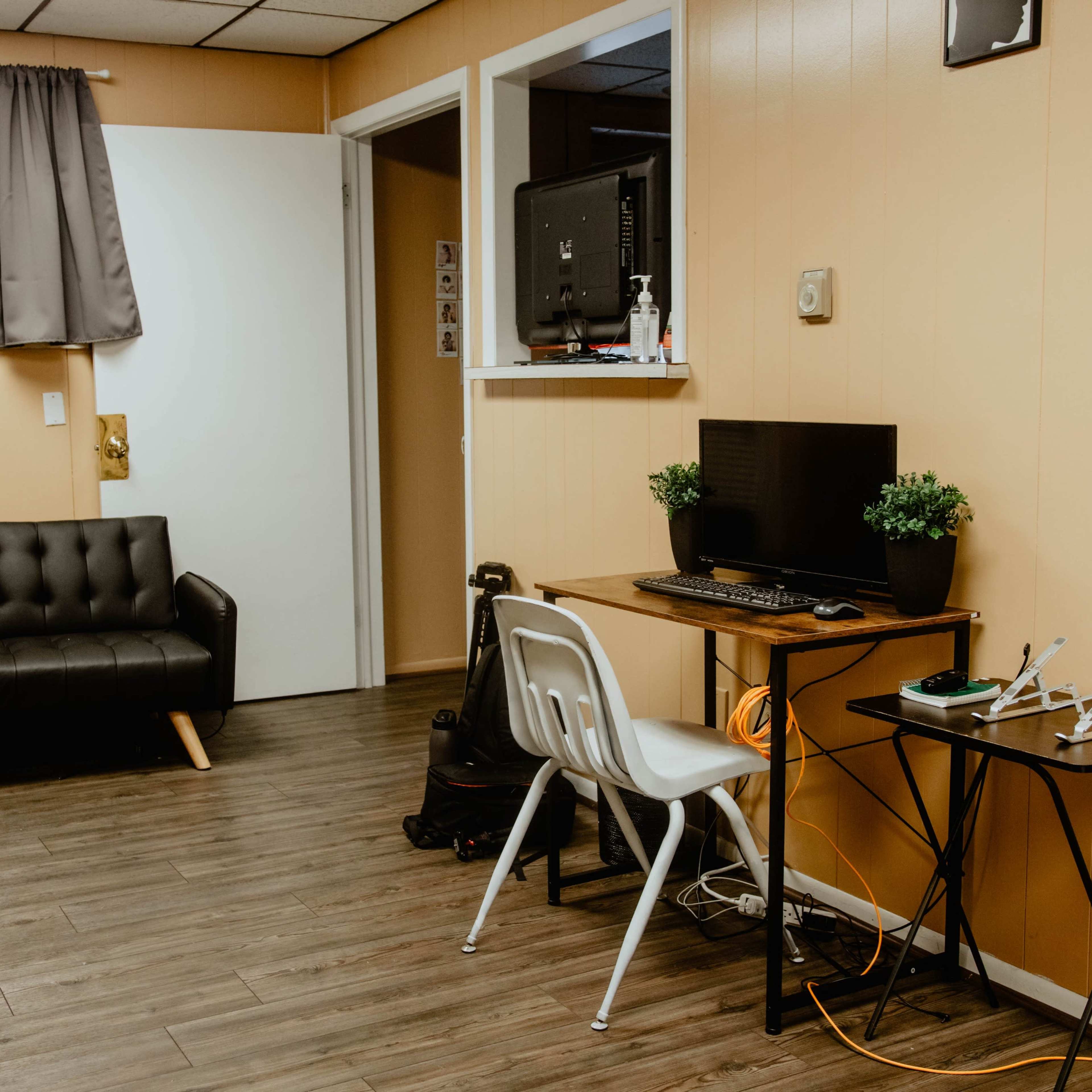 A small room with a black couch, a desk with a computer setup, a white chair, and a door leading to another area, all featuring light brown walls and wooden flooring.