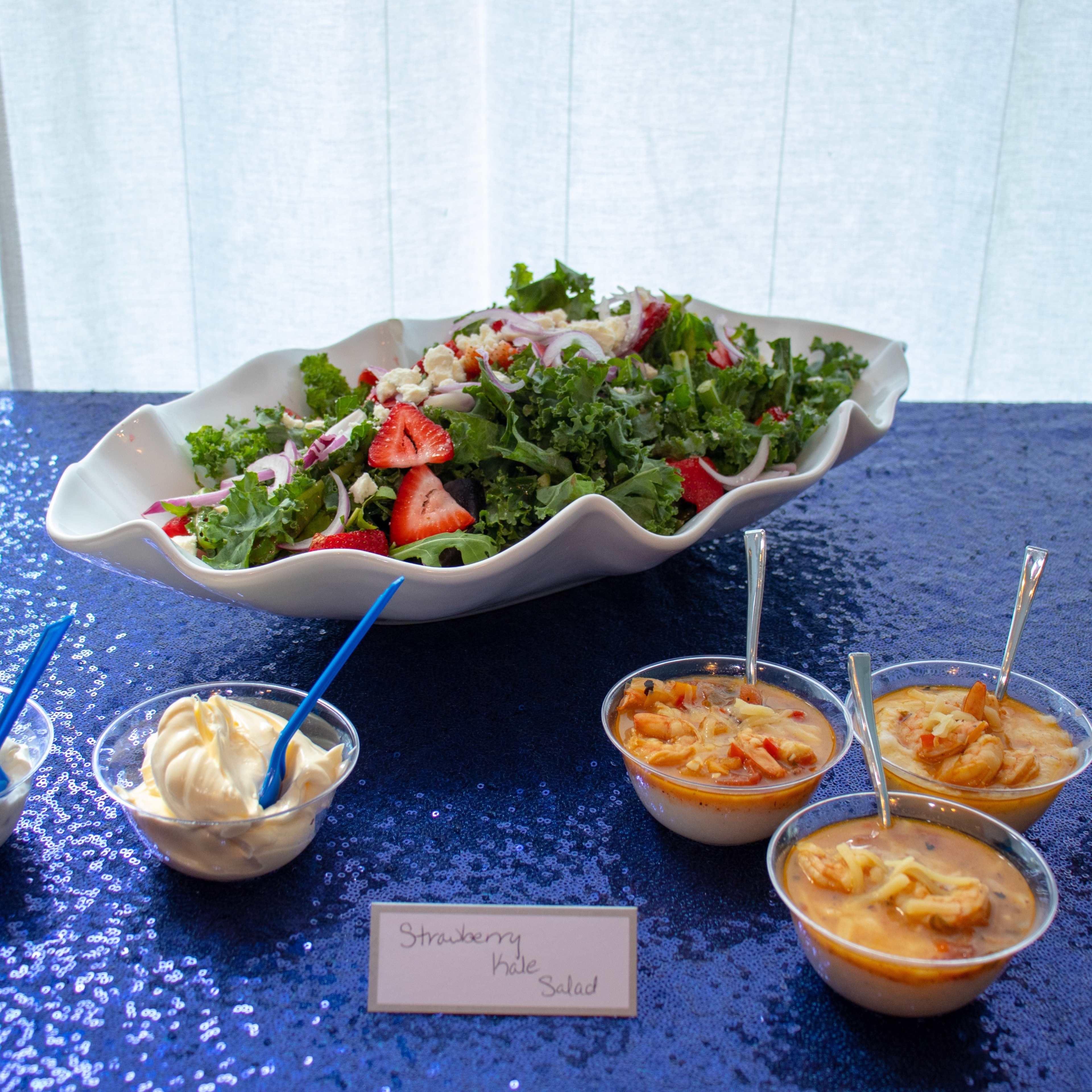 A large bowl of mixed greens and strawberries is placed beside six small cups of various sauces and a label reading "Strawberry Salad."