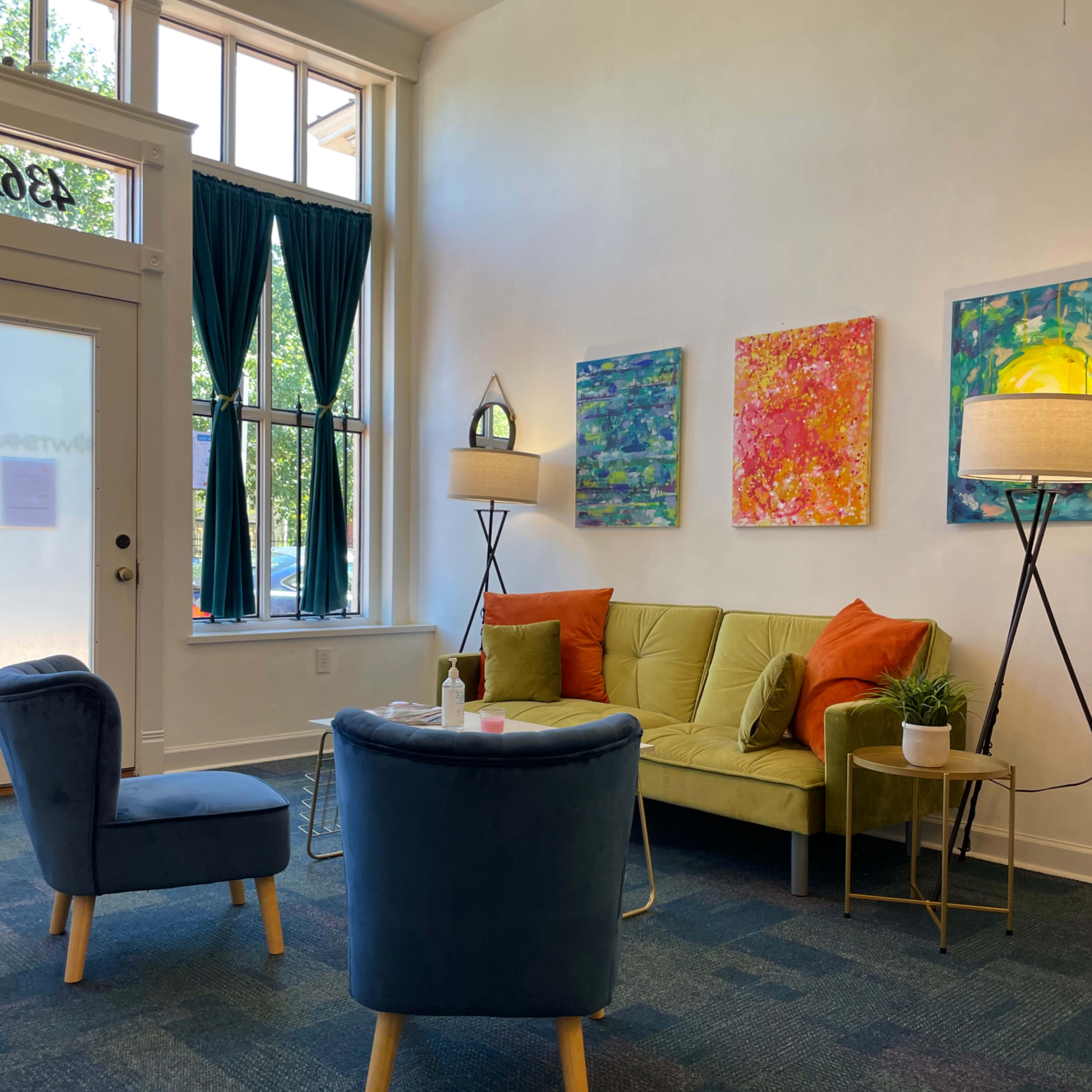 A waiting area with a green sofa, colorful wall art, and two lamps, accompanied by a blue armchair and a coffee table.