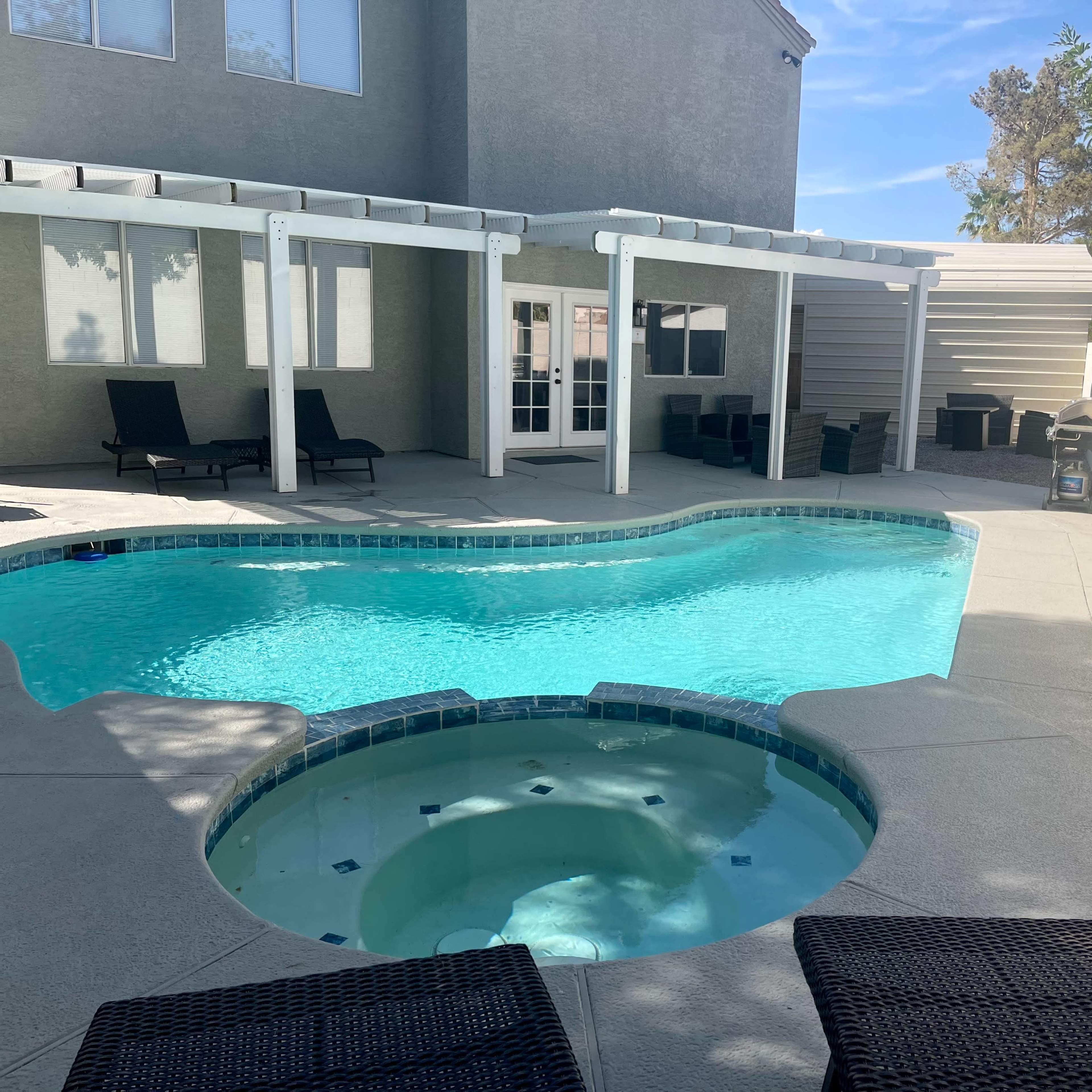 A swimming pool with an attached spa, surrounded by a concrete patio and shaded seating areas beside a house.