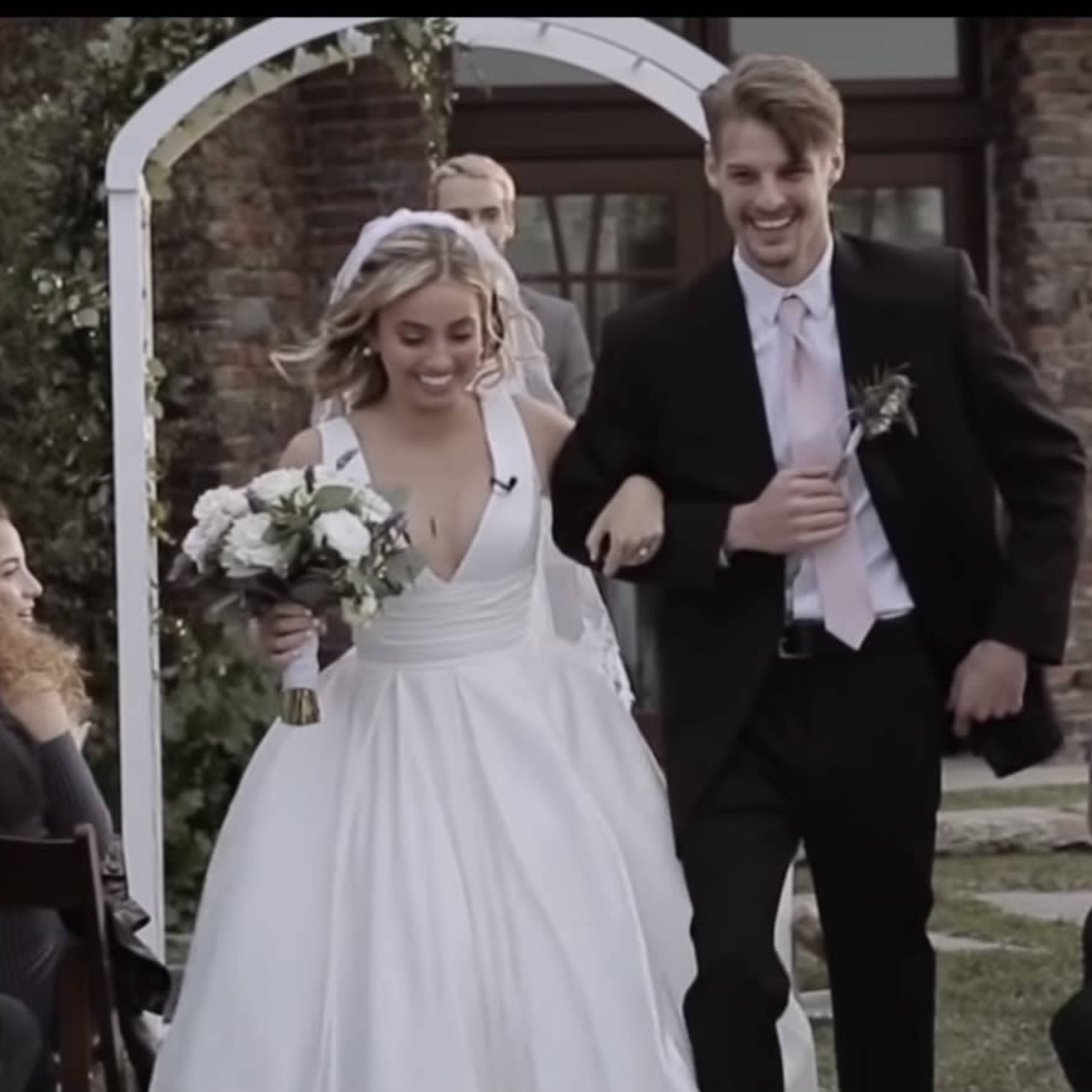 A bride and groom walk down the aisle together, smiling, as guests cheer and applaud in a garden setting.