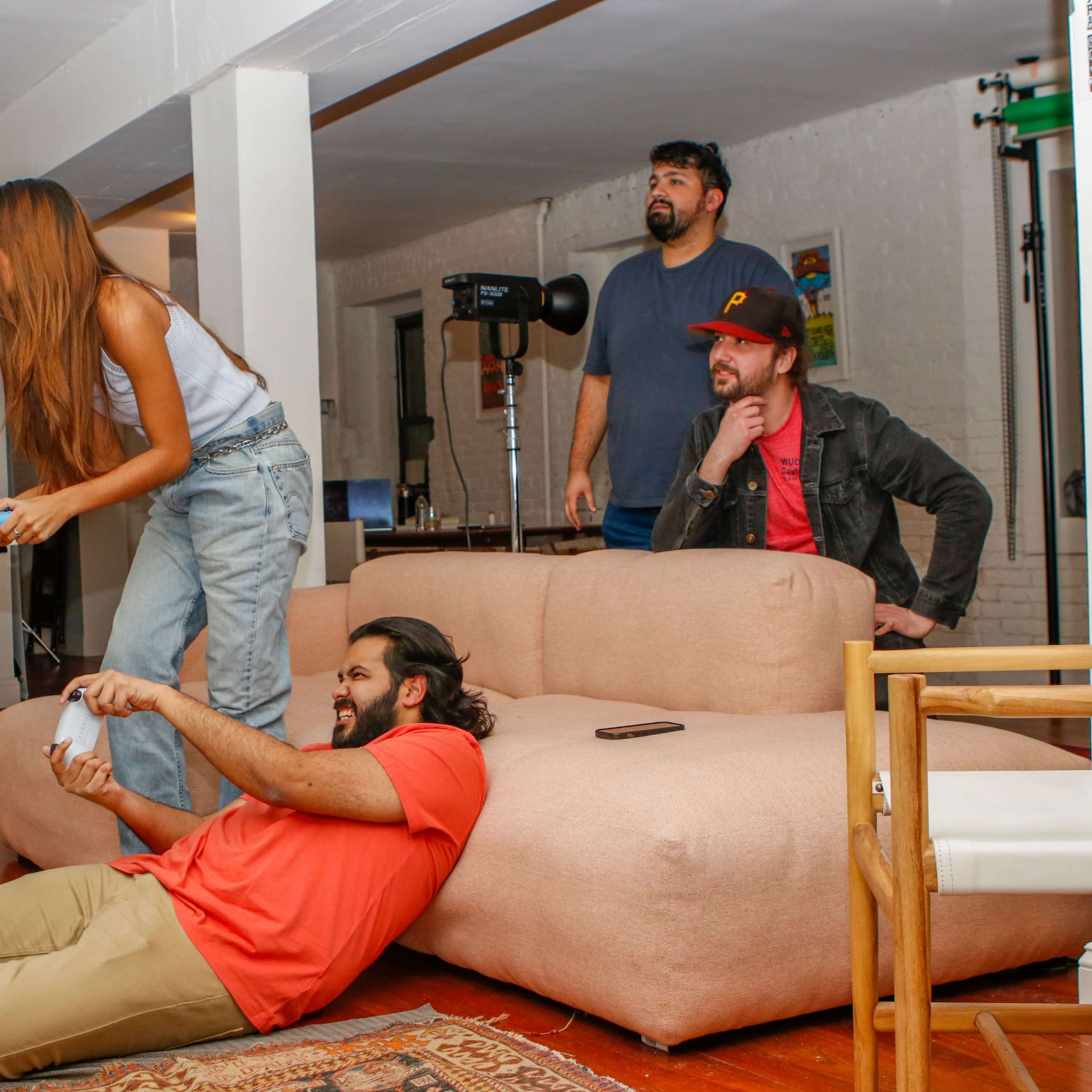 A group of four people is gathered in a modern living space, with one person lying on the floor playing a video game while the others watch from a couch and a nearby chair.