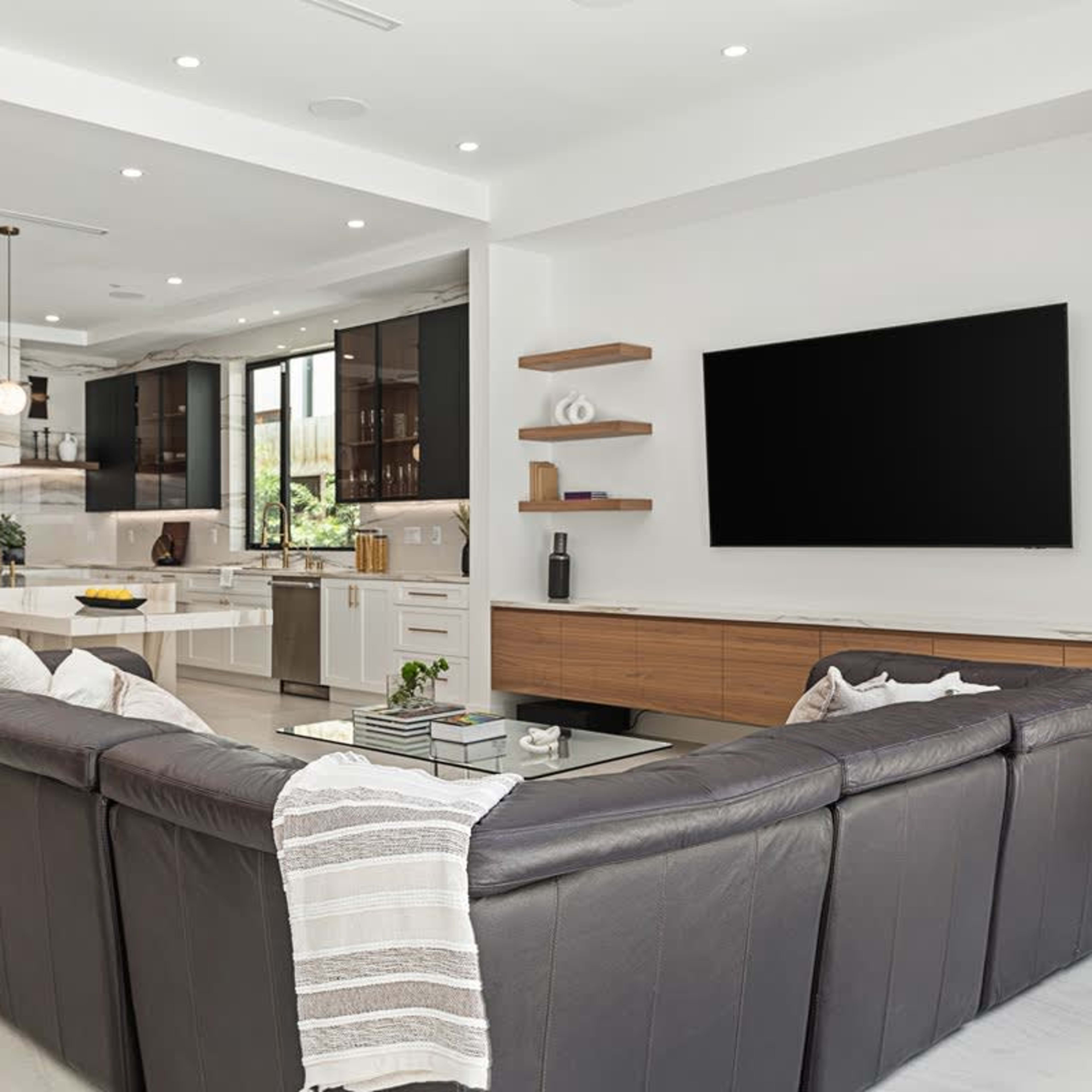 A modern living room with a sectional sofa, a coffee table, and a flat-screen television mounted on the wall, adjacent to an open kitchen with sleek cabinetry.