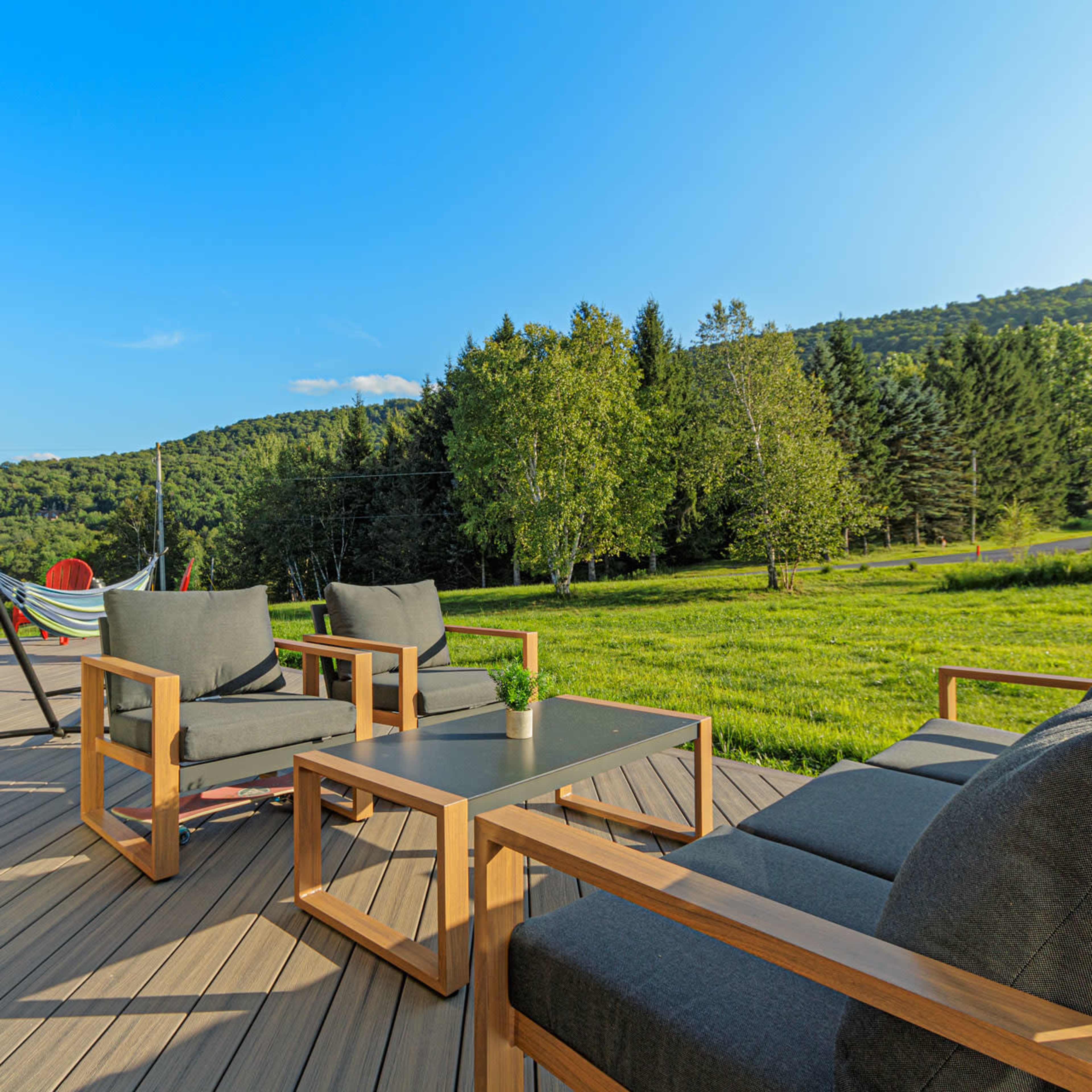 A patio area featuring wooden furniture, a hammock, and a red umbrella, overlooking a grassy field and wooded hills.