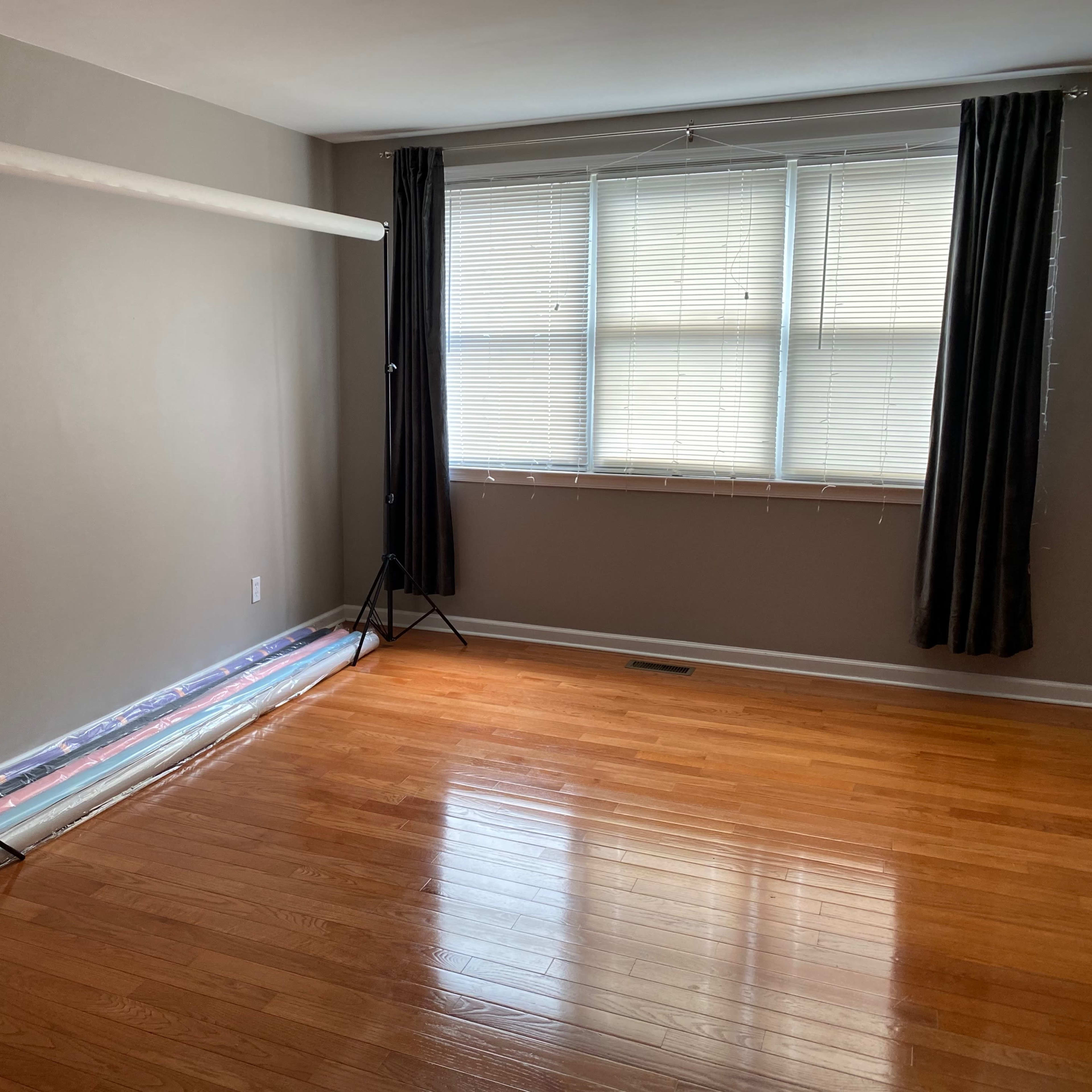 An empty room with polished wooden flooring, a backdrop stand with white fabric, and a roll of backdrop material on the floor near a large window with horizontal blinds.