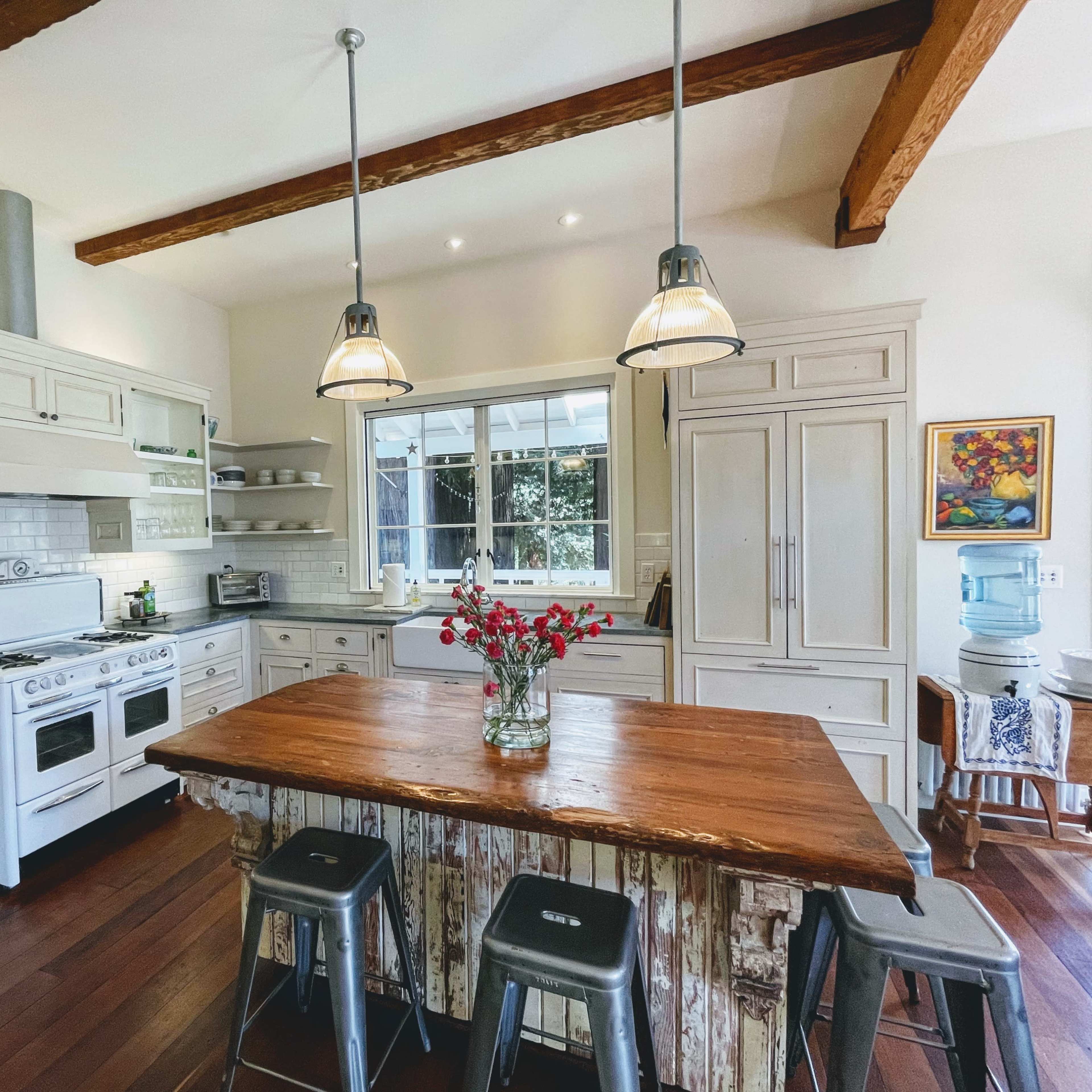 A kitchen features a large wooden island, white cabinets, and pendant lights hanging from exposed beams.
