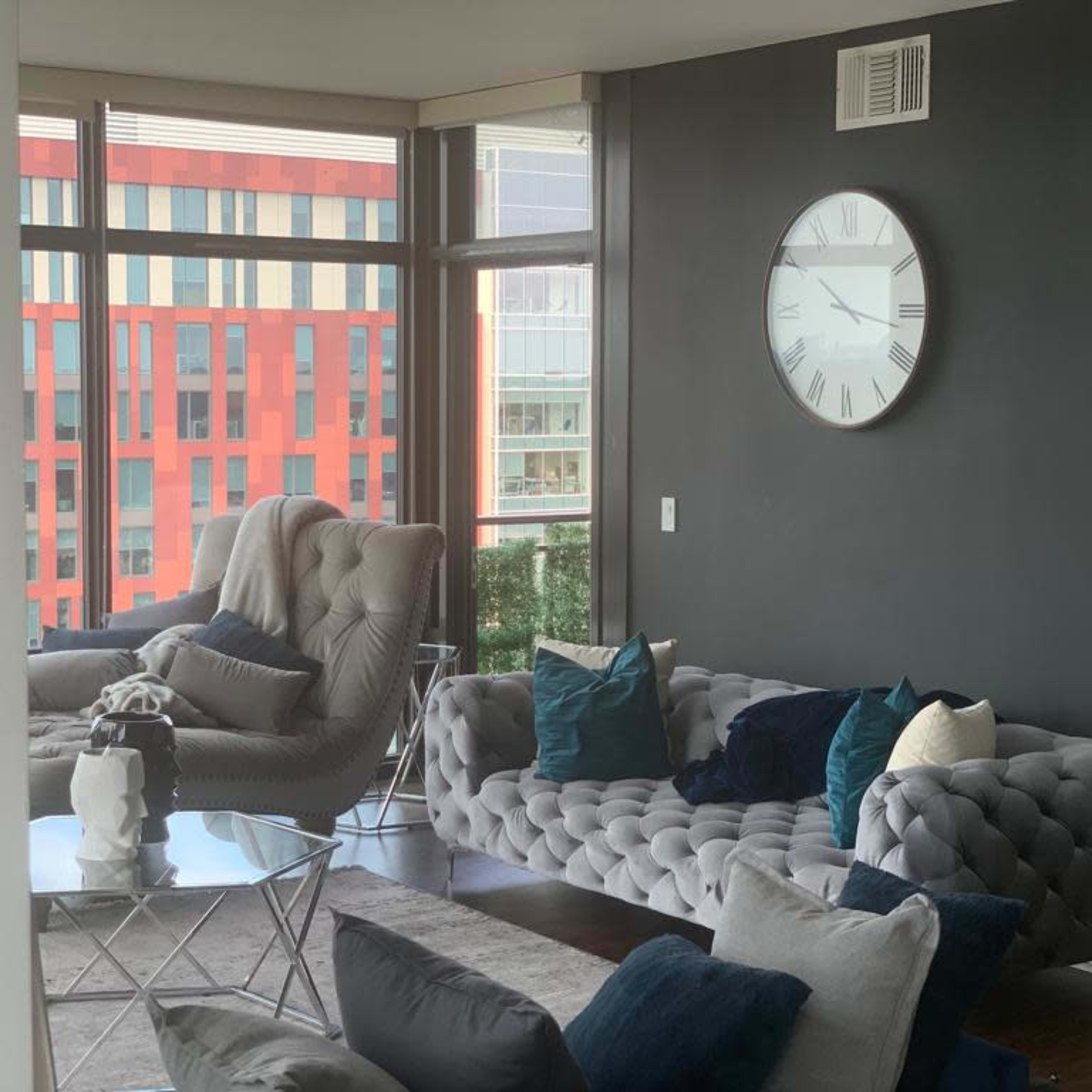 A modern living room with a gray sectional sofa, a glass coffee table, and large windows overlooking a colorful building.