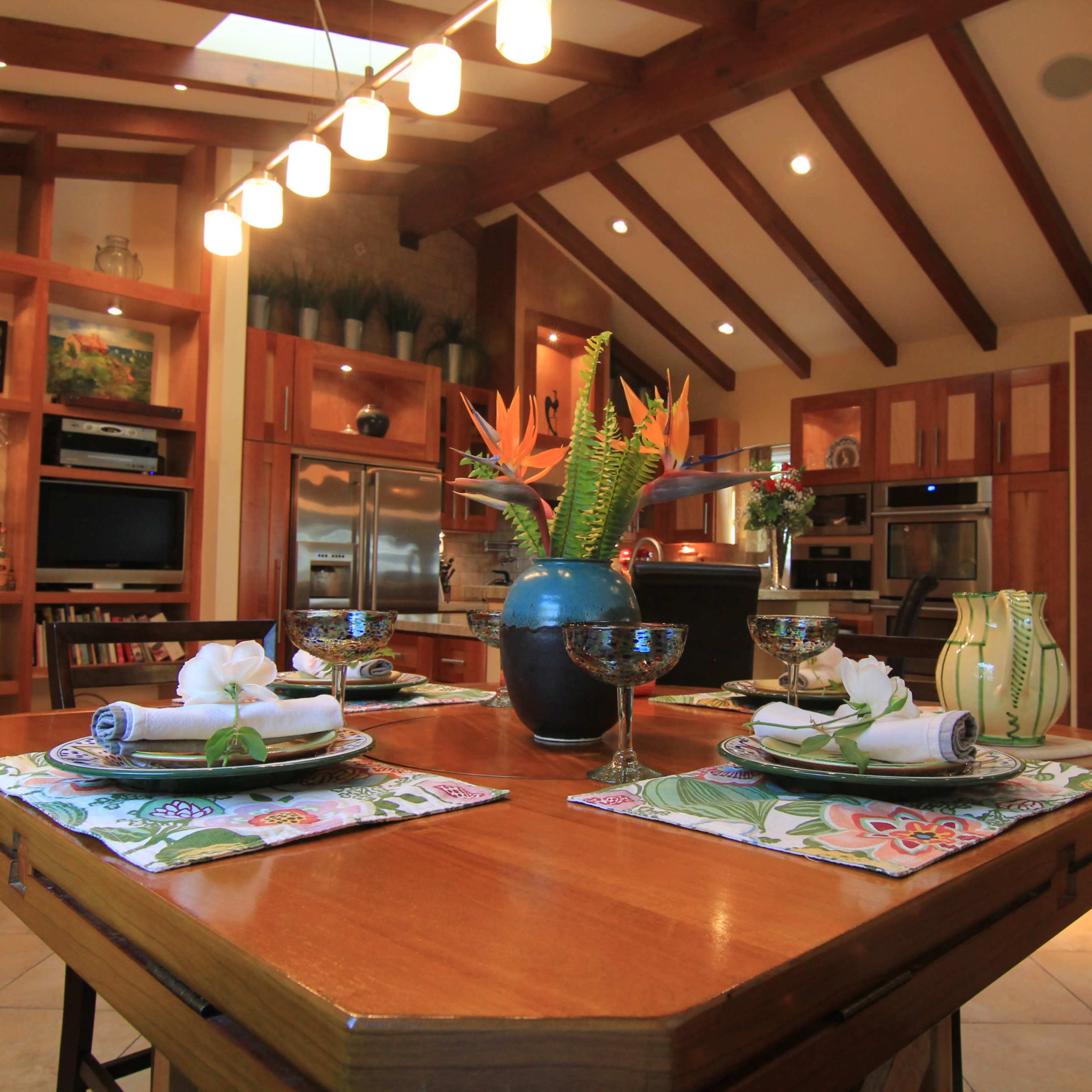 A dining table is set with plates and flowers in a bright kitchen featuring wooden beams and built-in shelves.