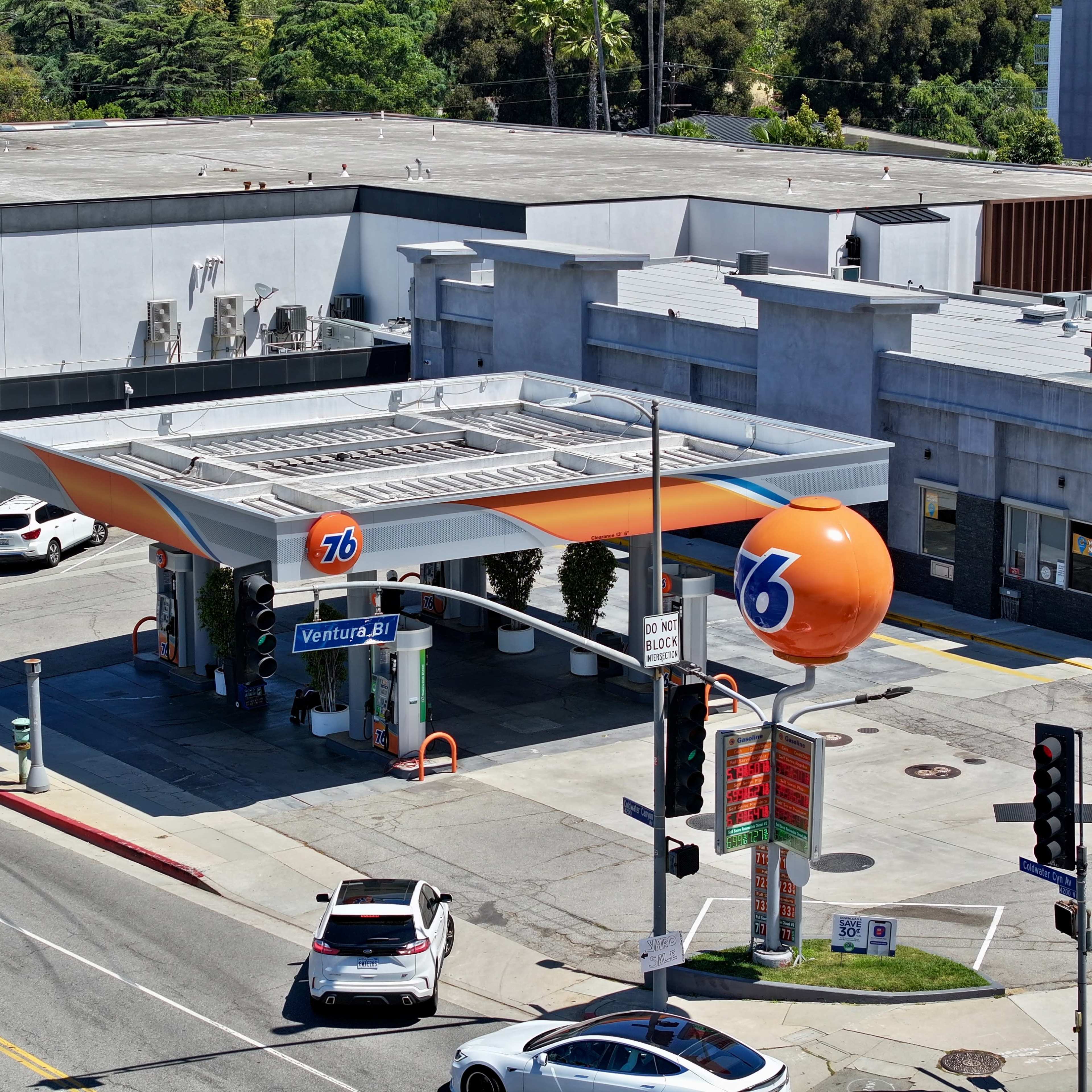 A 76 gas station with a distinctive orange logo is situated at a street intersection, surrounded by buildings and parked cars.