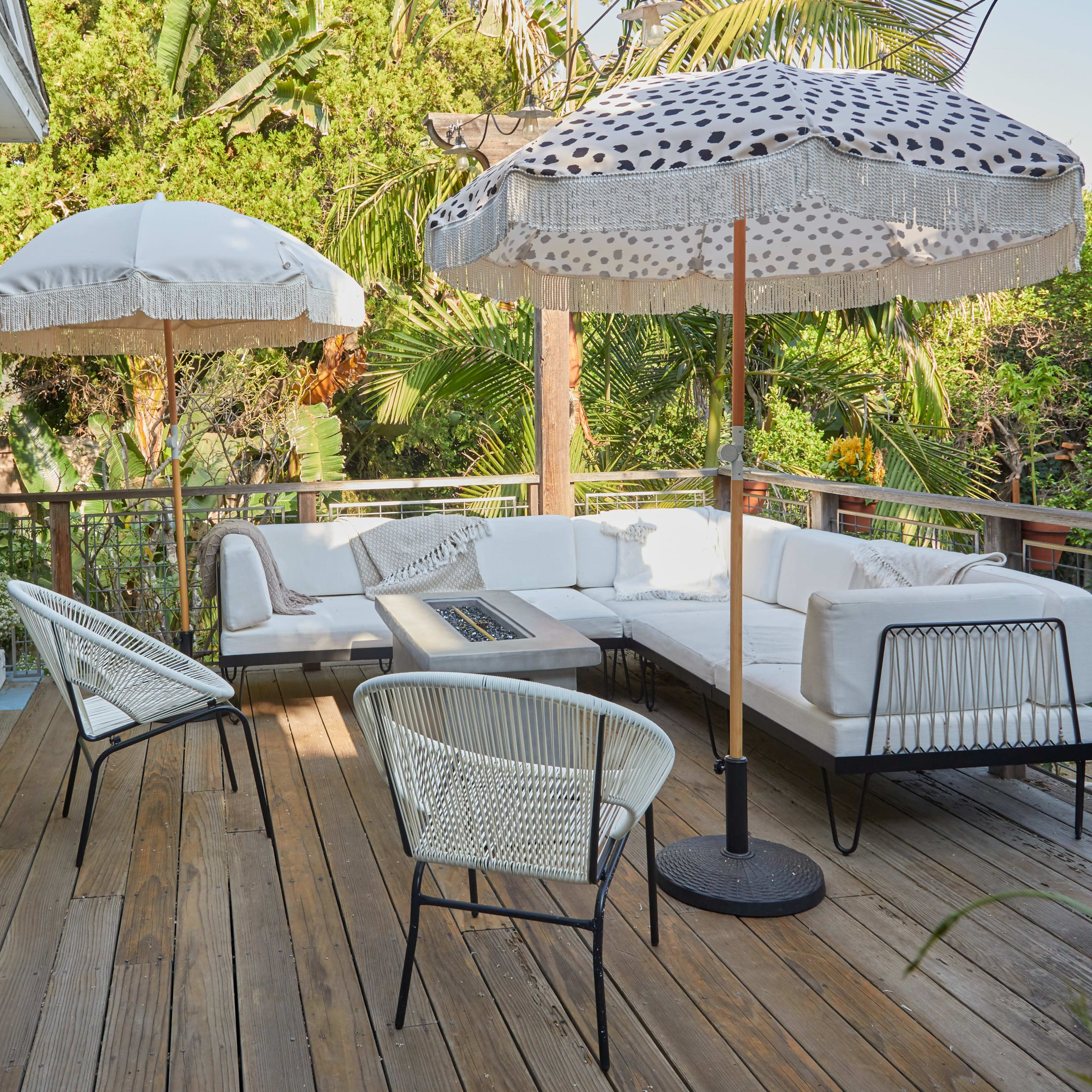 A deck features a sectional sofa and four chairs, shaded by two large umbrellas surrounded by lush greenery.