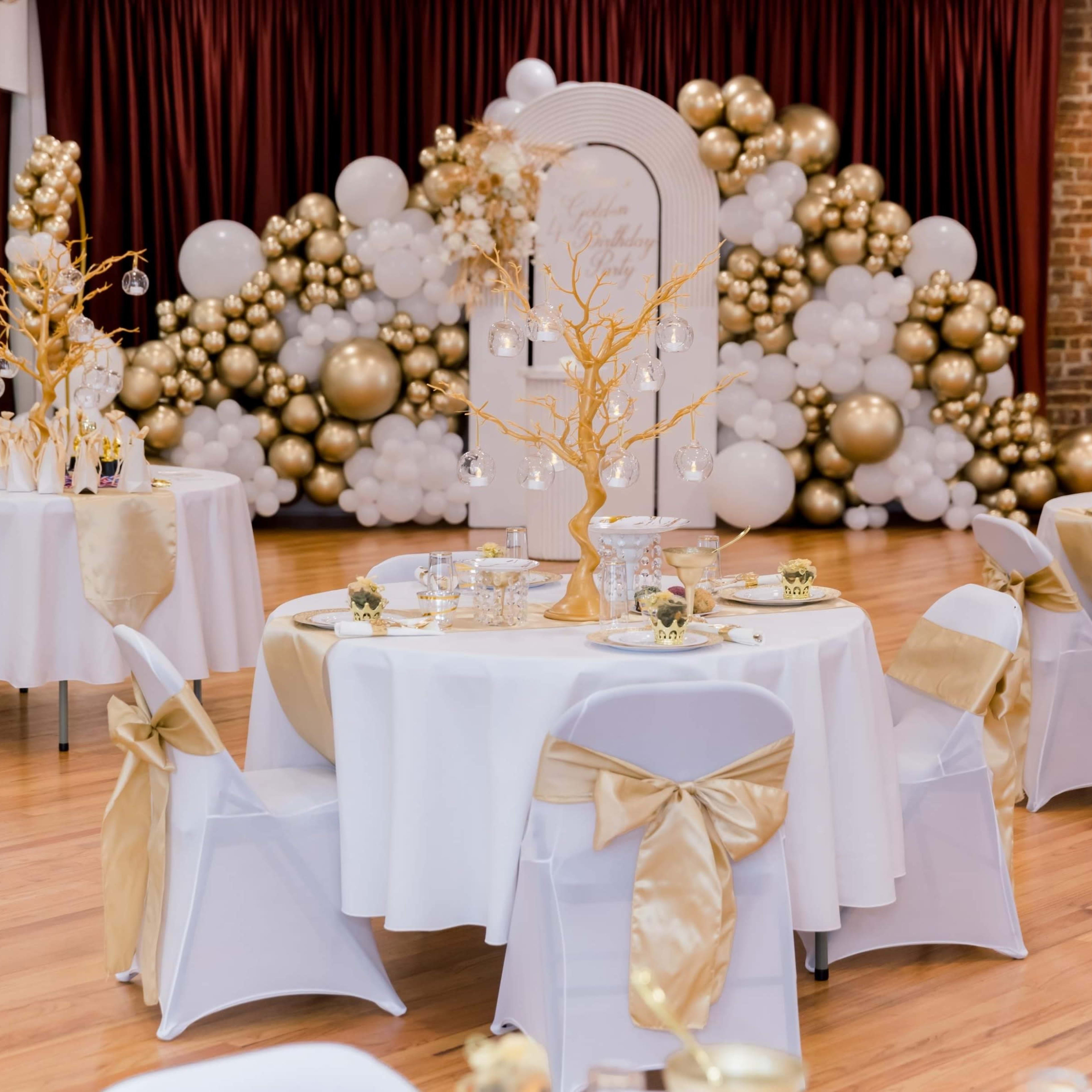 A decorated event space featuring round tables with white tablecloths and gold accents, surrounded by a backdrop of gold and white balloon arrangements.