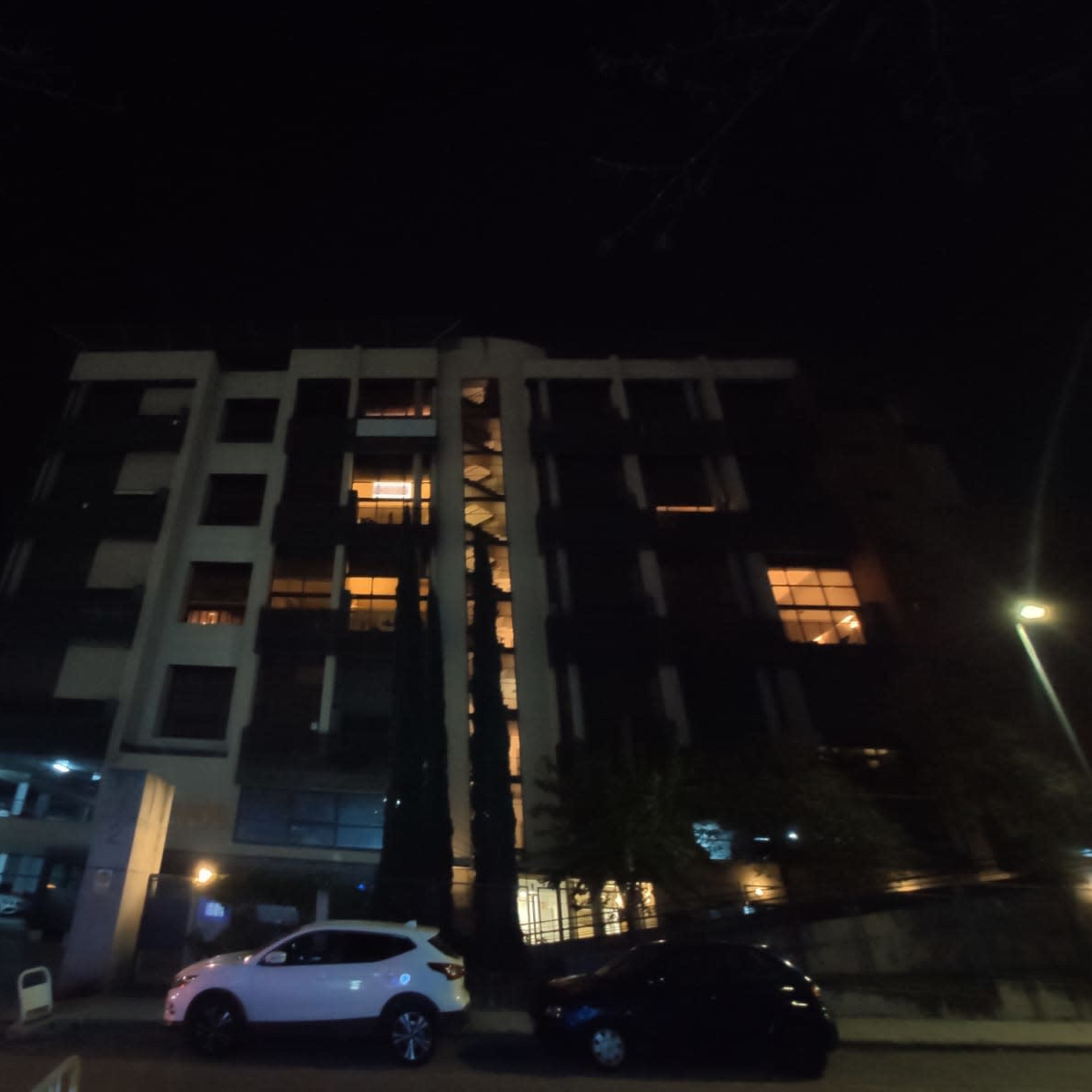 A multi-story residential building at night with illuminated windows and parked cars in the foreground.